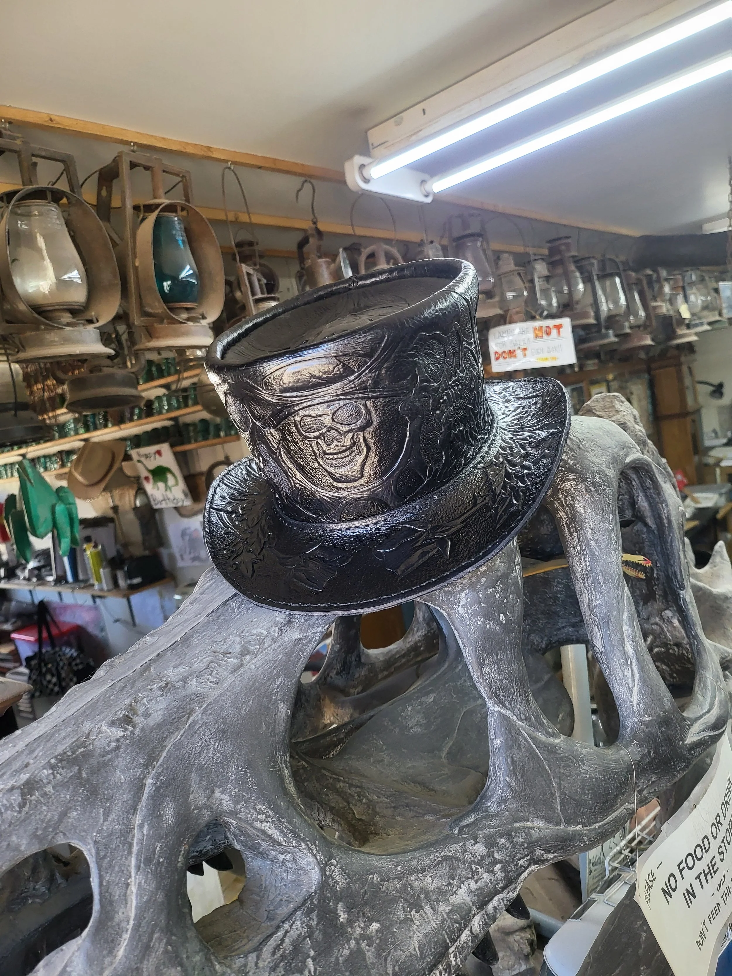Decorative black top hat with a skull and mustache design, placed on a rustic wooden bench in a store with vintage lanterns hanging on the wall behind.