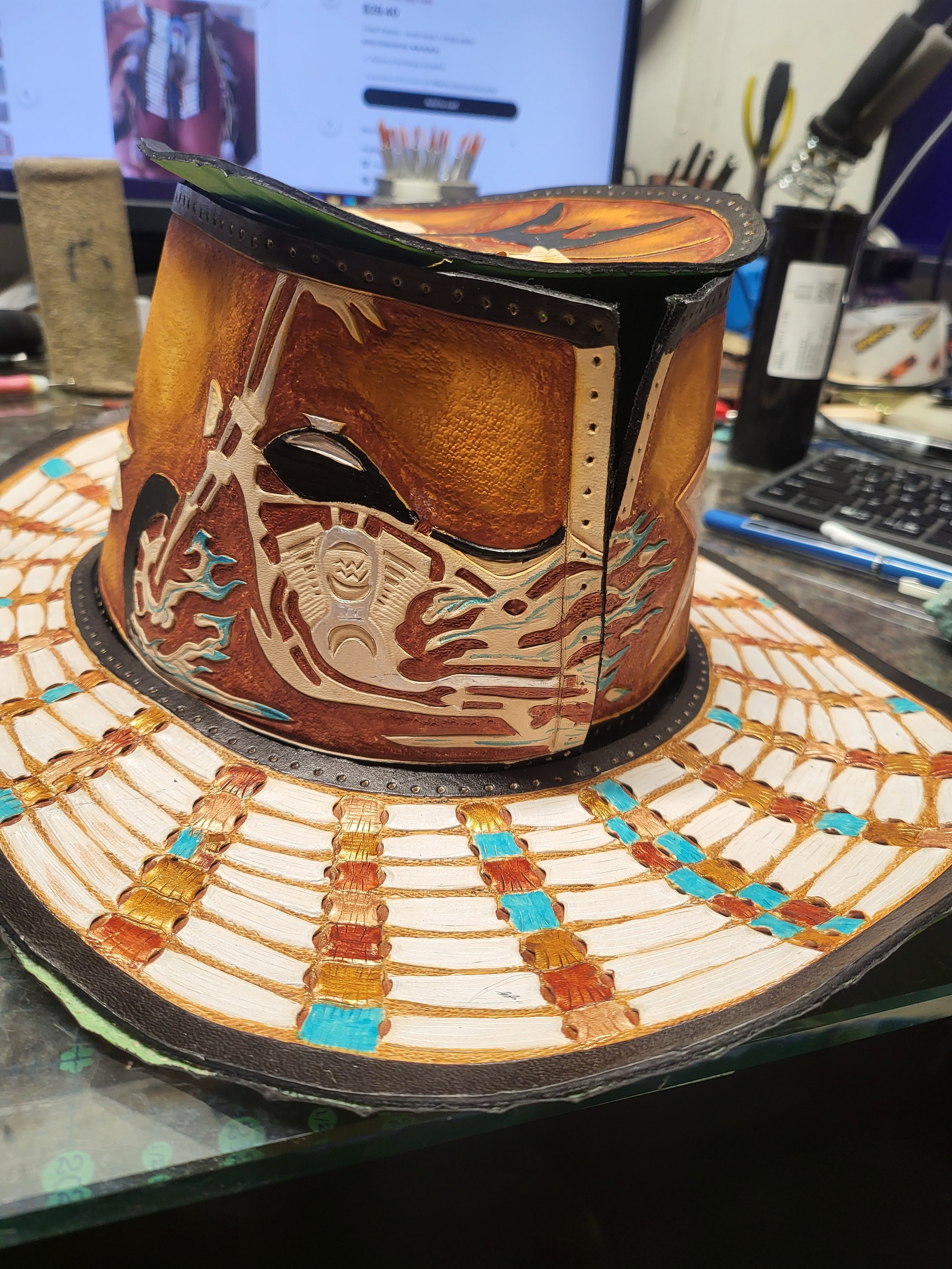A handmade leather and painted cowboy hat with a Star Wars Mandalorian helmet design on the front, placed on a cluttered desk with tools and a computer monitor in the background.