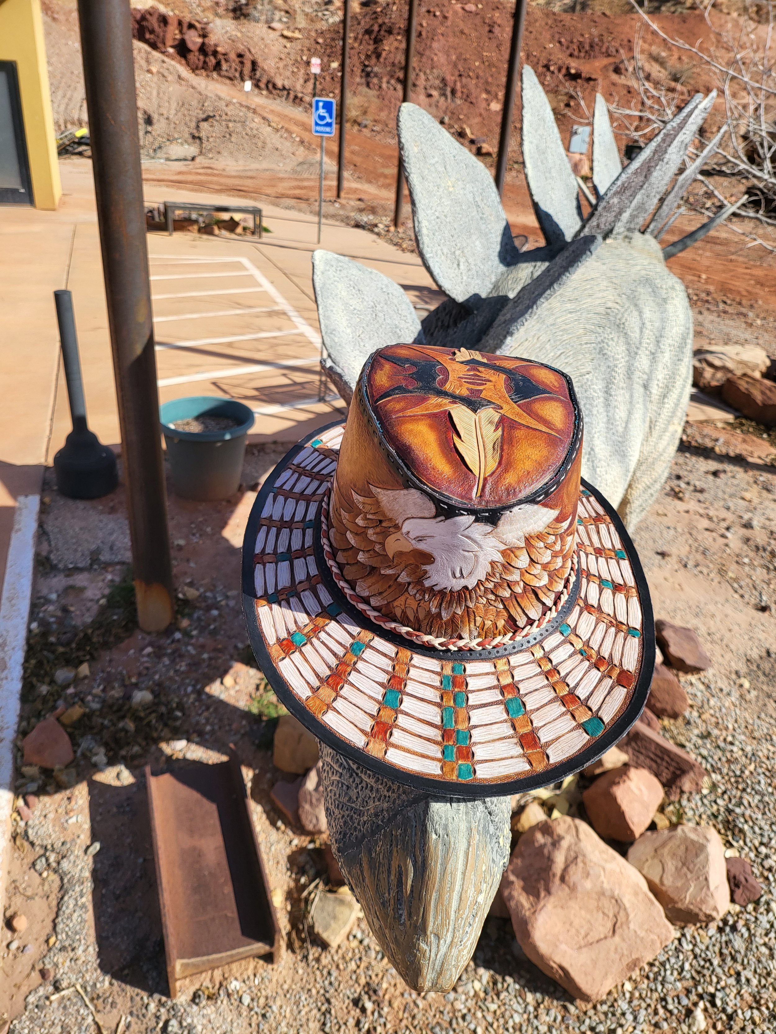 Decorative wide-brimmed hat with a beaded design placed on top of a carved wooden stake at an outdoor area with a stone sculpture of a rabbit, a handicap parking sign, and mountain landscape in the background.