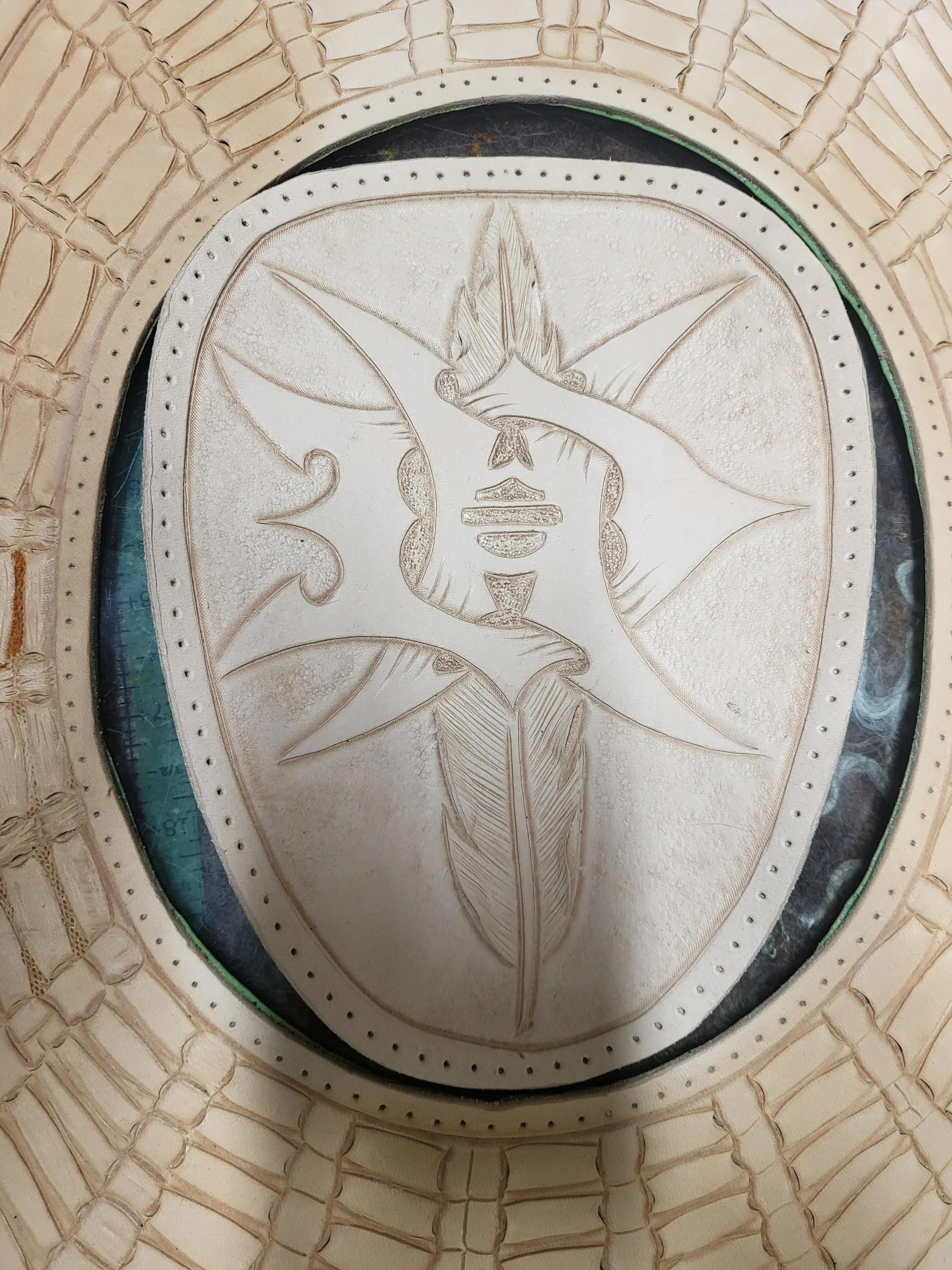 Close-up of a carved tribal or Indigenous art piece depicting a face within a star-shaped design, surrounded by a decorative oval border with dotted patterns, mounted on a woven basket or similar craft item.