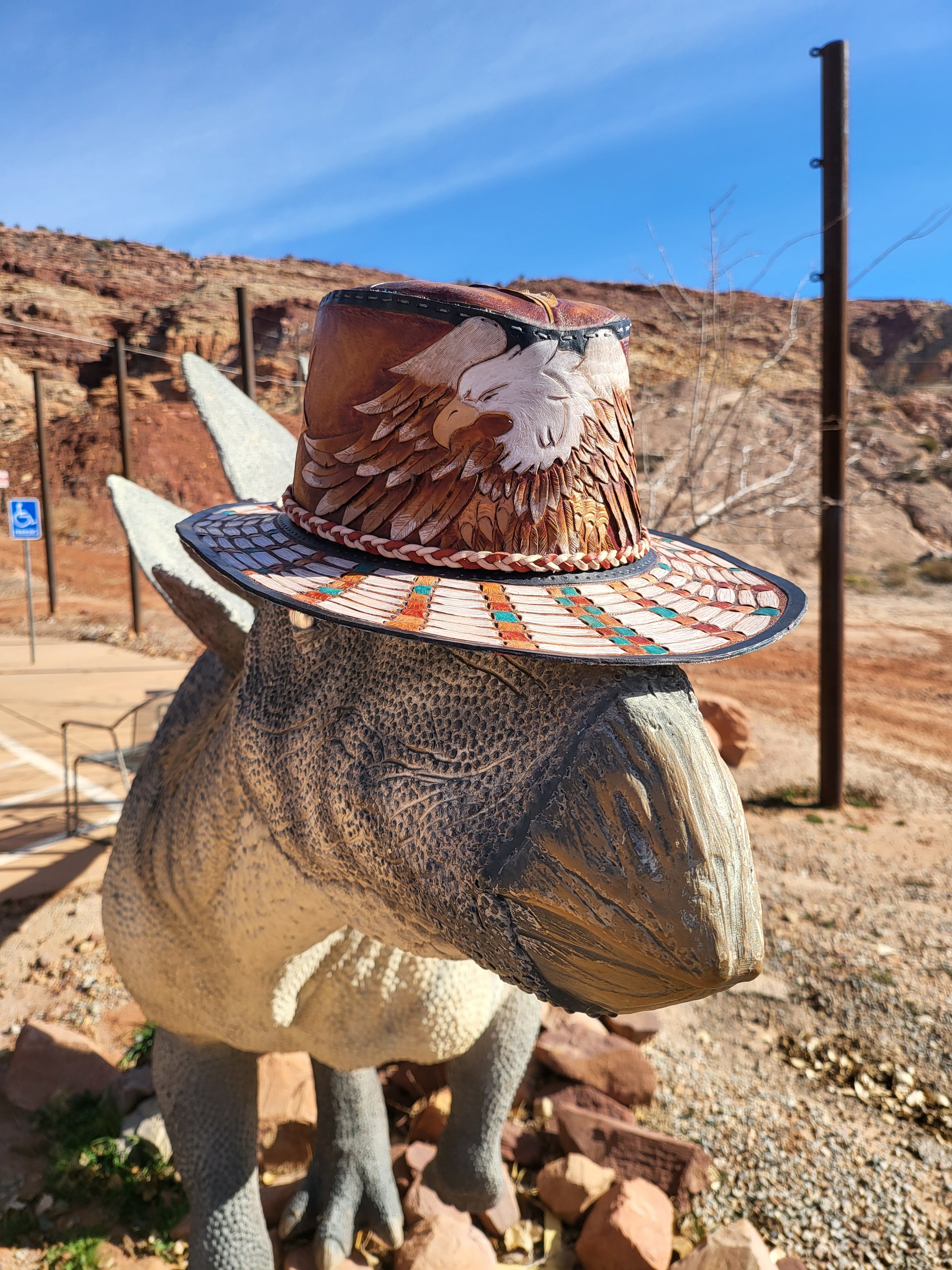 A dinosaur statue, likely a velociraptor, with a straw hat featuring an eagle painted on it, outdoors against a desert landscape with red rocks and a clear blue sky.