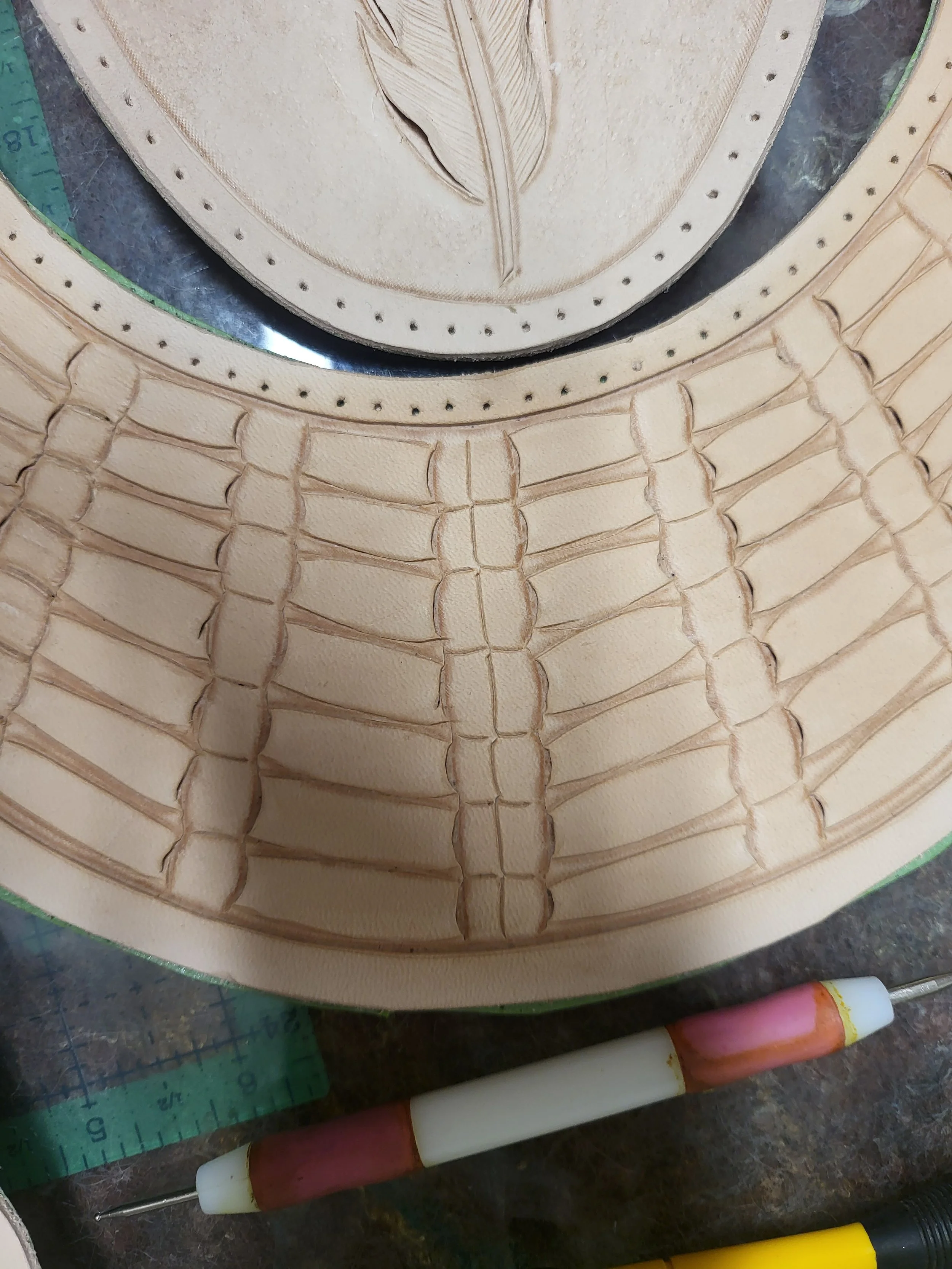 A close-up of a leatherwork project showing stitched details on a tan leather piece with a carved feather design. Nearby are a green ruler, a pink and white handle tool, and a yellow marker.