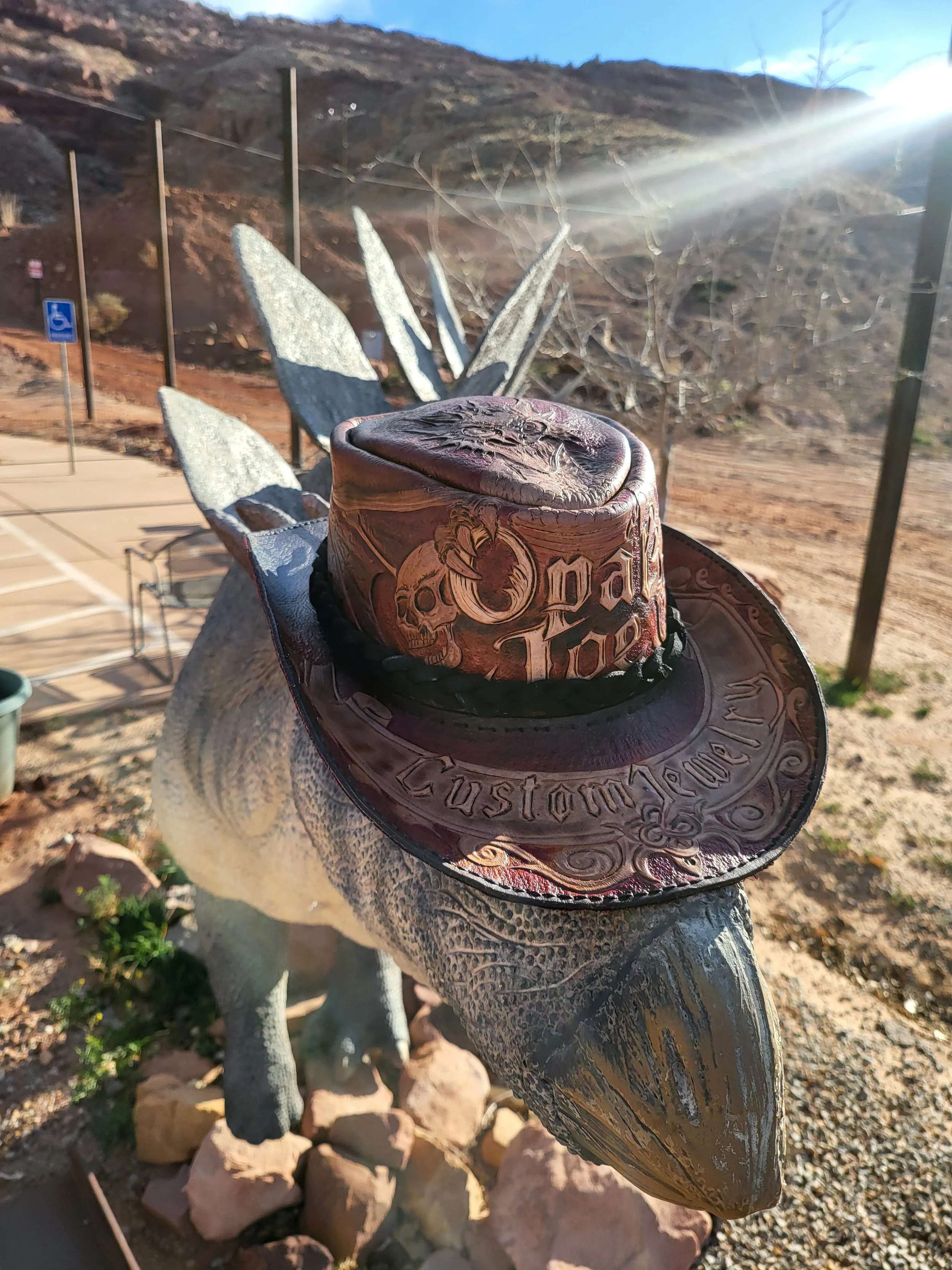 A cowboy hat with decorative skull and text is placed on a statue of a dinosaur outside in a desert landscape.