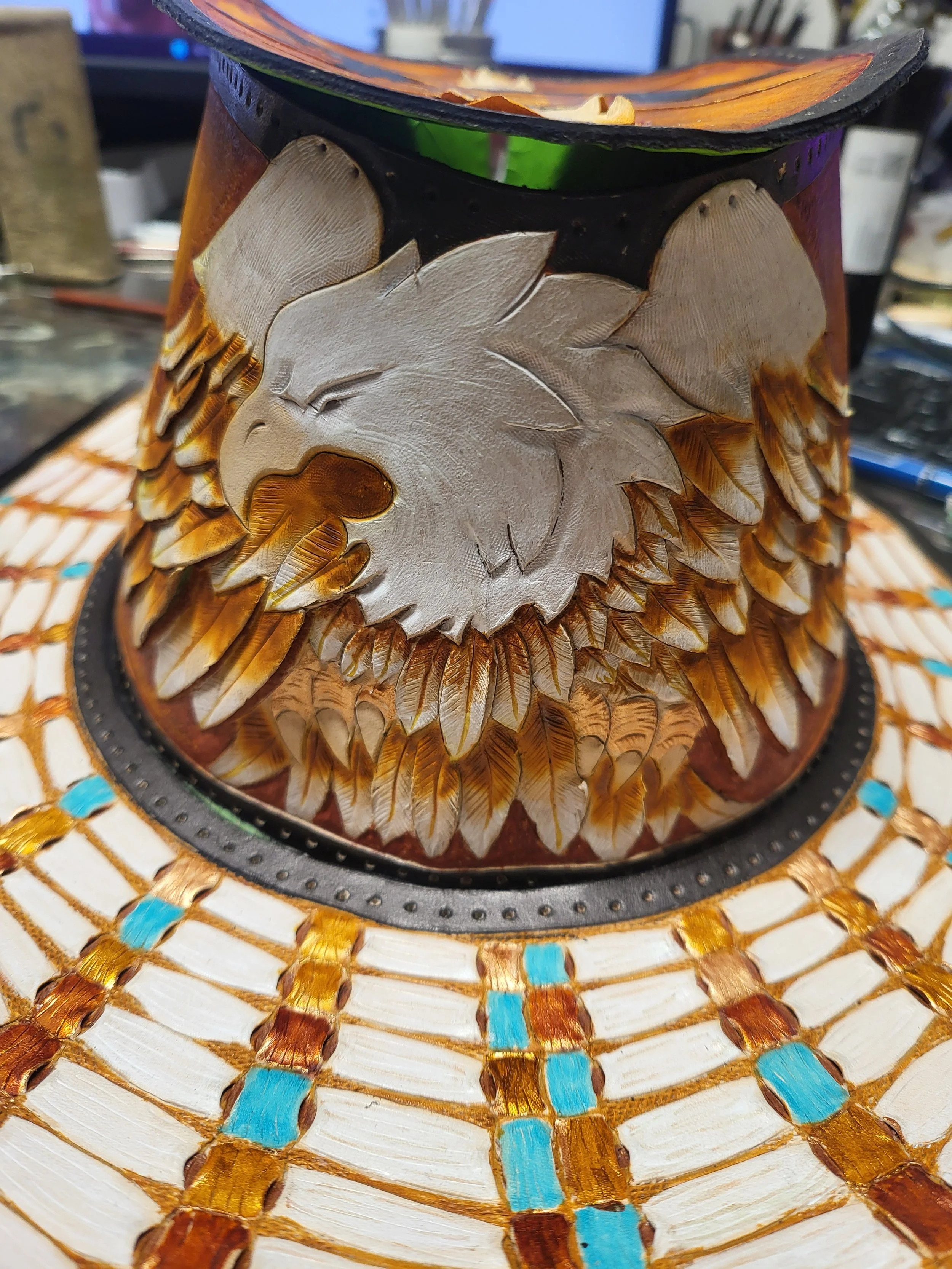 Close-up of a decorated cowboy hat with a carved horse head emblem, featuring feather-like designs and a woven pattern with blue accents.