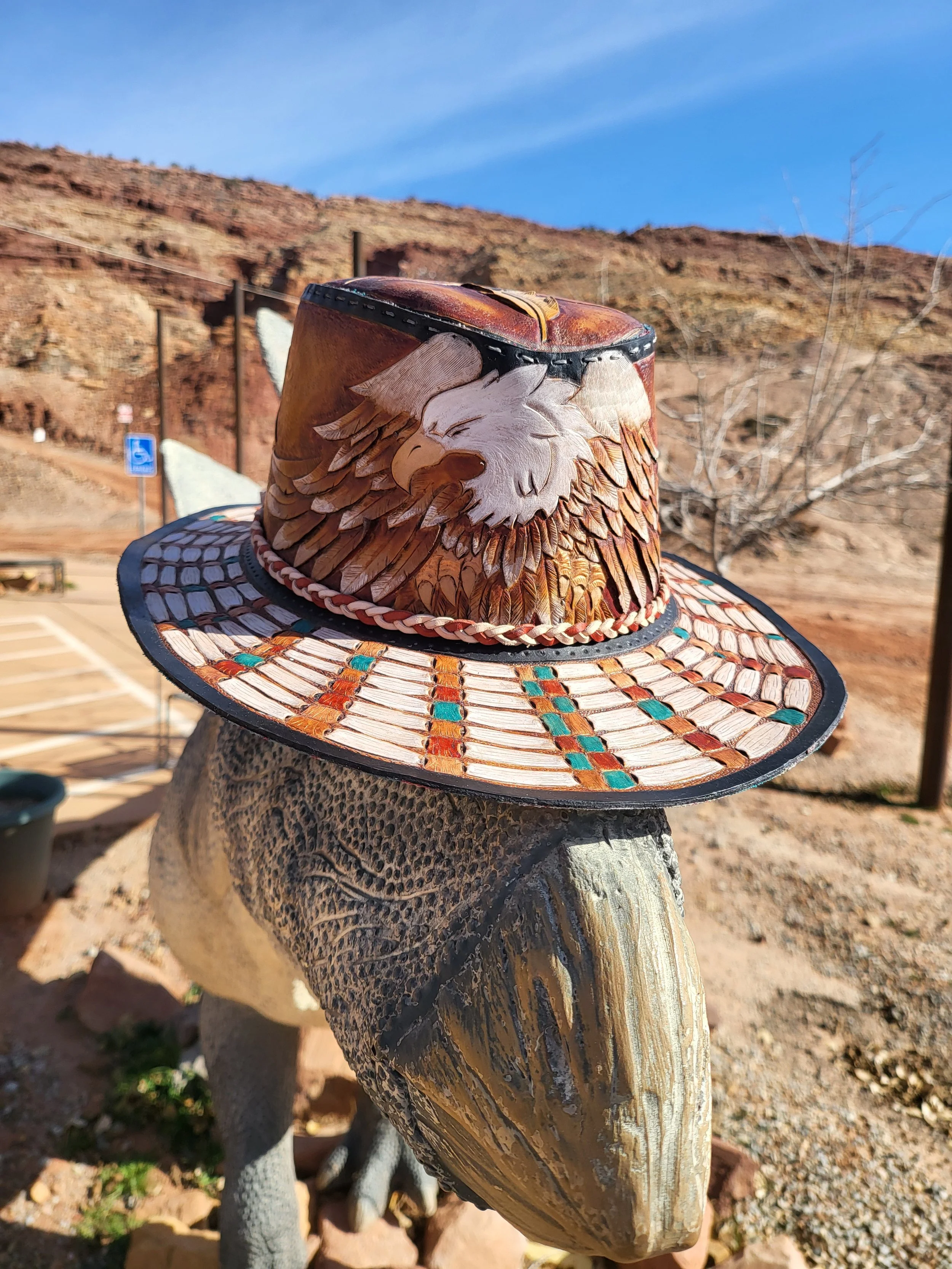 Decorative felt hat with an eagle design on the front, placed on a dinosaur sculpture in a desert landscape with red rock hills in the background.