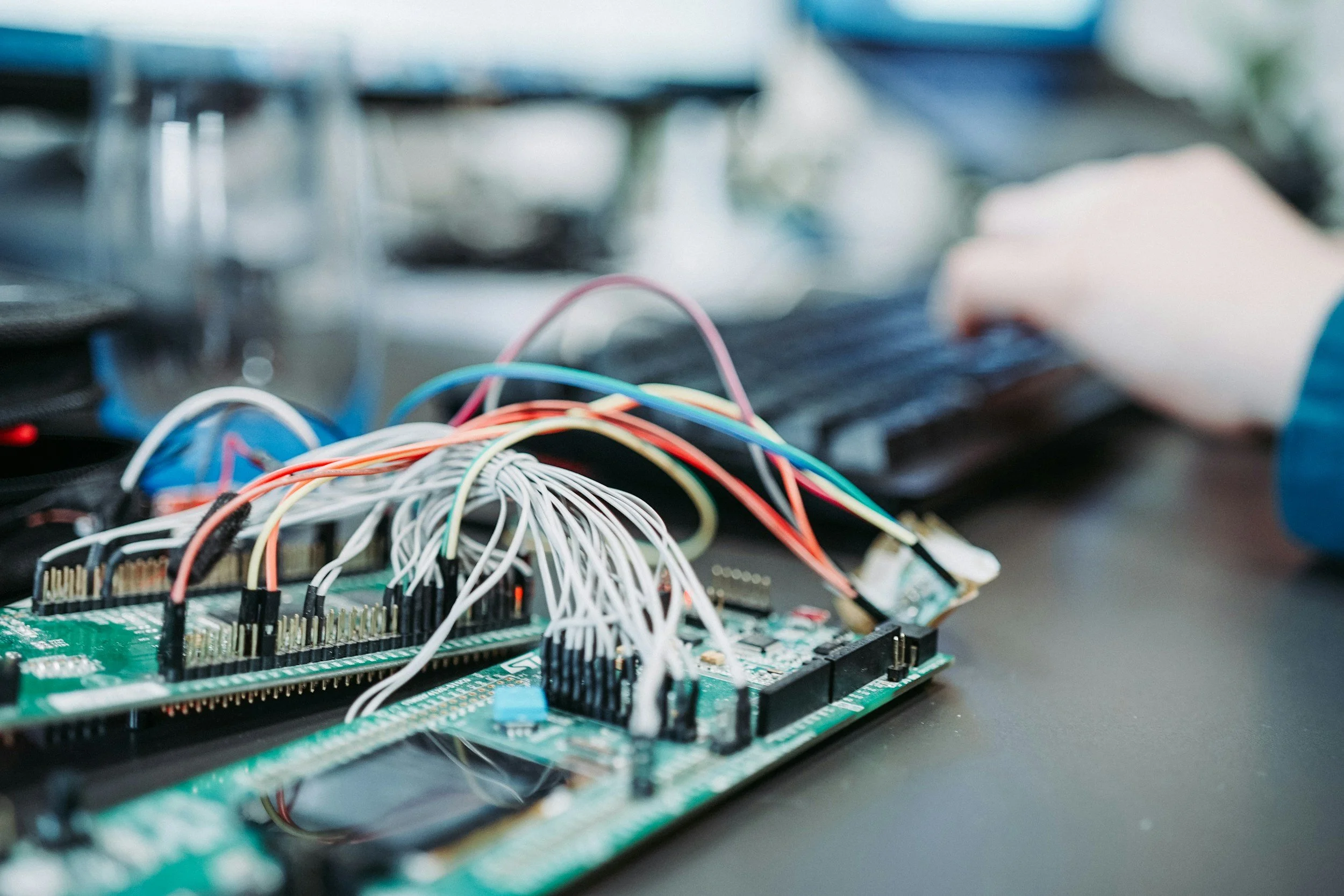 Close-up view of a green circuit board with numerous multicolored wires connected to it, and a person's hand working on a keyboard in the background.
