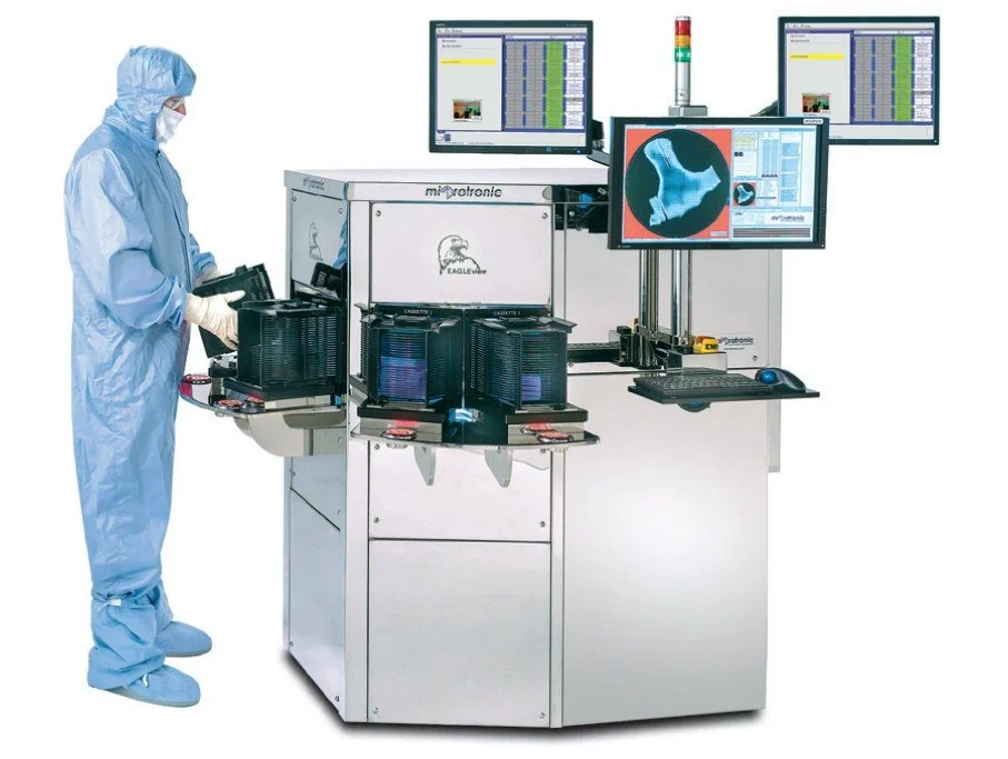 A scientist in a blue cleanroom suit working with a microplate reader instrument connected to multiple monitors displaying scientific data and images.