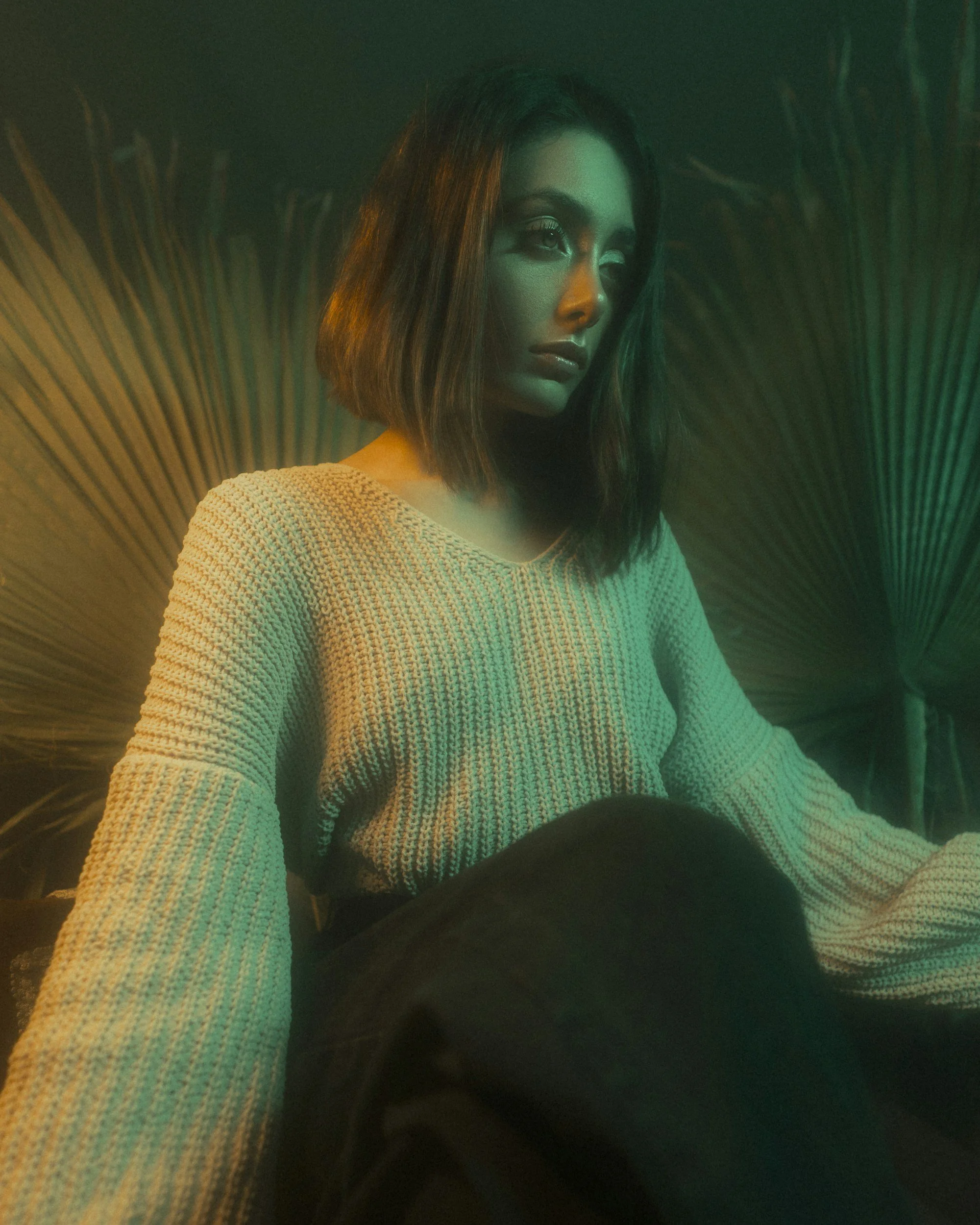 A woman with shoulder-length dark hair and makeup, wearing a cream-colored knitted sweater, sitting in front of large green palm leaves, illuminated by green and yellow lighting.