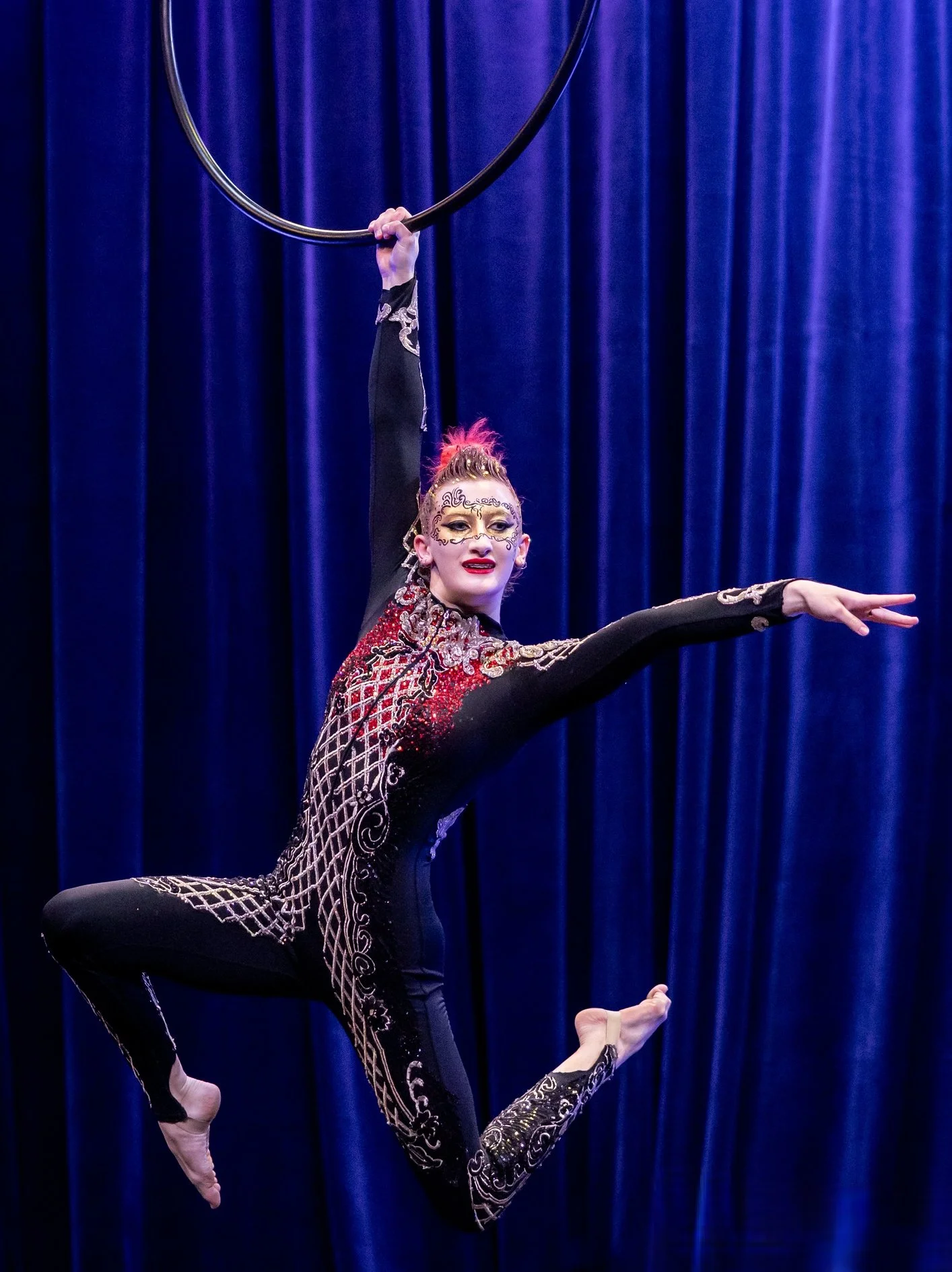A female acrobat performing an aerial pose holding a hoop above her head with her right hand while balancing in a split with her left leg extended forward and right leg bent behind her. She is dressed in a black costume with intricate gold and silver embroidery and face paint, performing on a stage with blue curtains in the background.