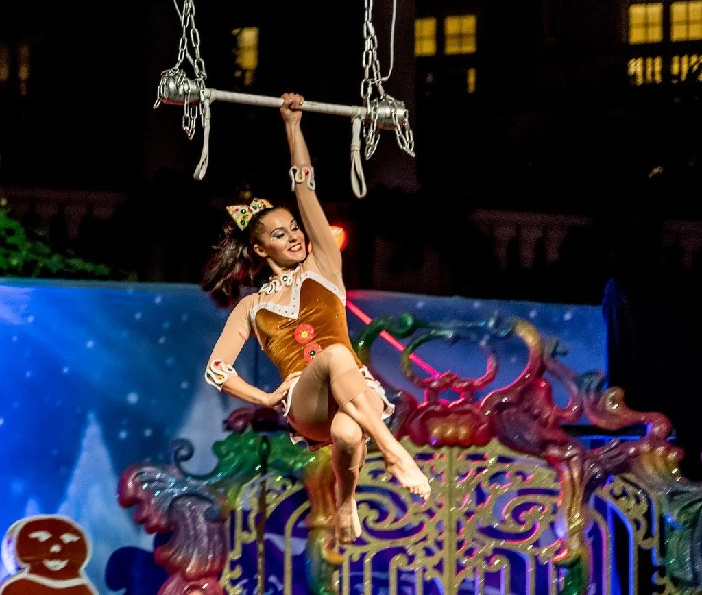 A woman in a vintage costume is swinging on a trapeze at night during a carnival or circus show, with colorful decorations and animated background.