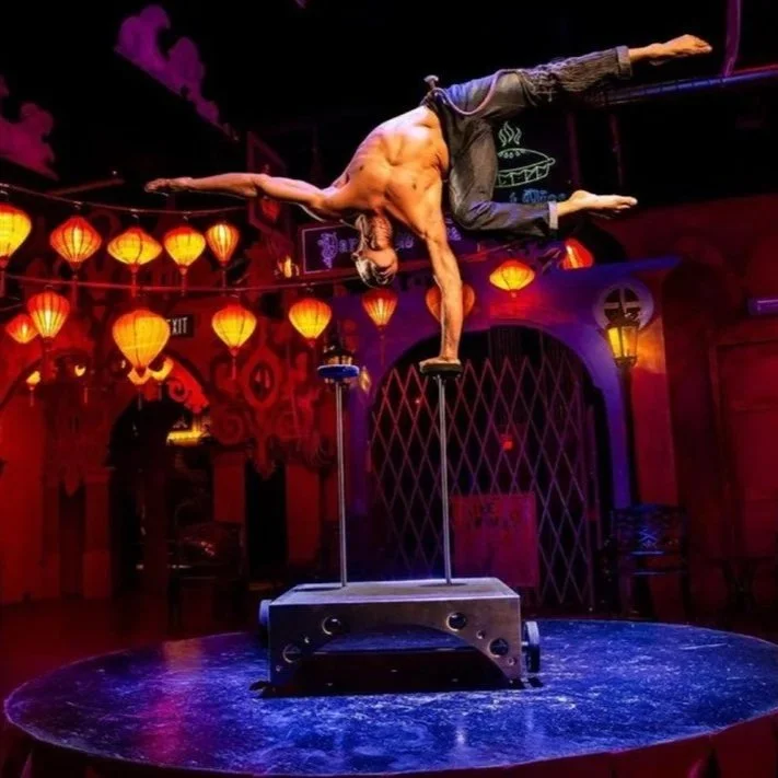 A performer balances one arm on a small platform on a circular stage, with a colorful Asian-themed lantern-lit background.