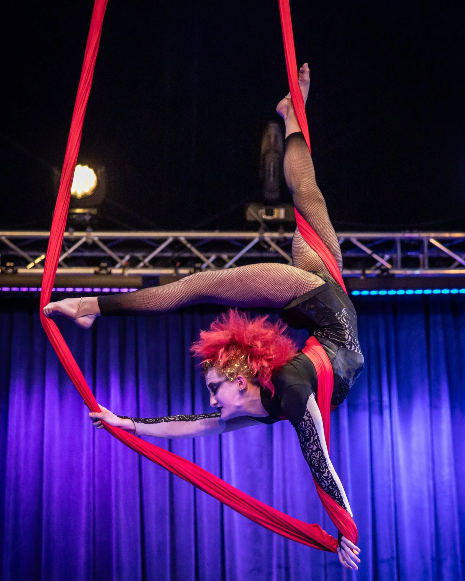 An aerial silk performer with bright red hair and face makeup, dressed in black and fishnet stockings, executing a pose on red silks with a dark stage background and purple curtains.