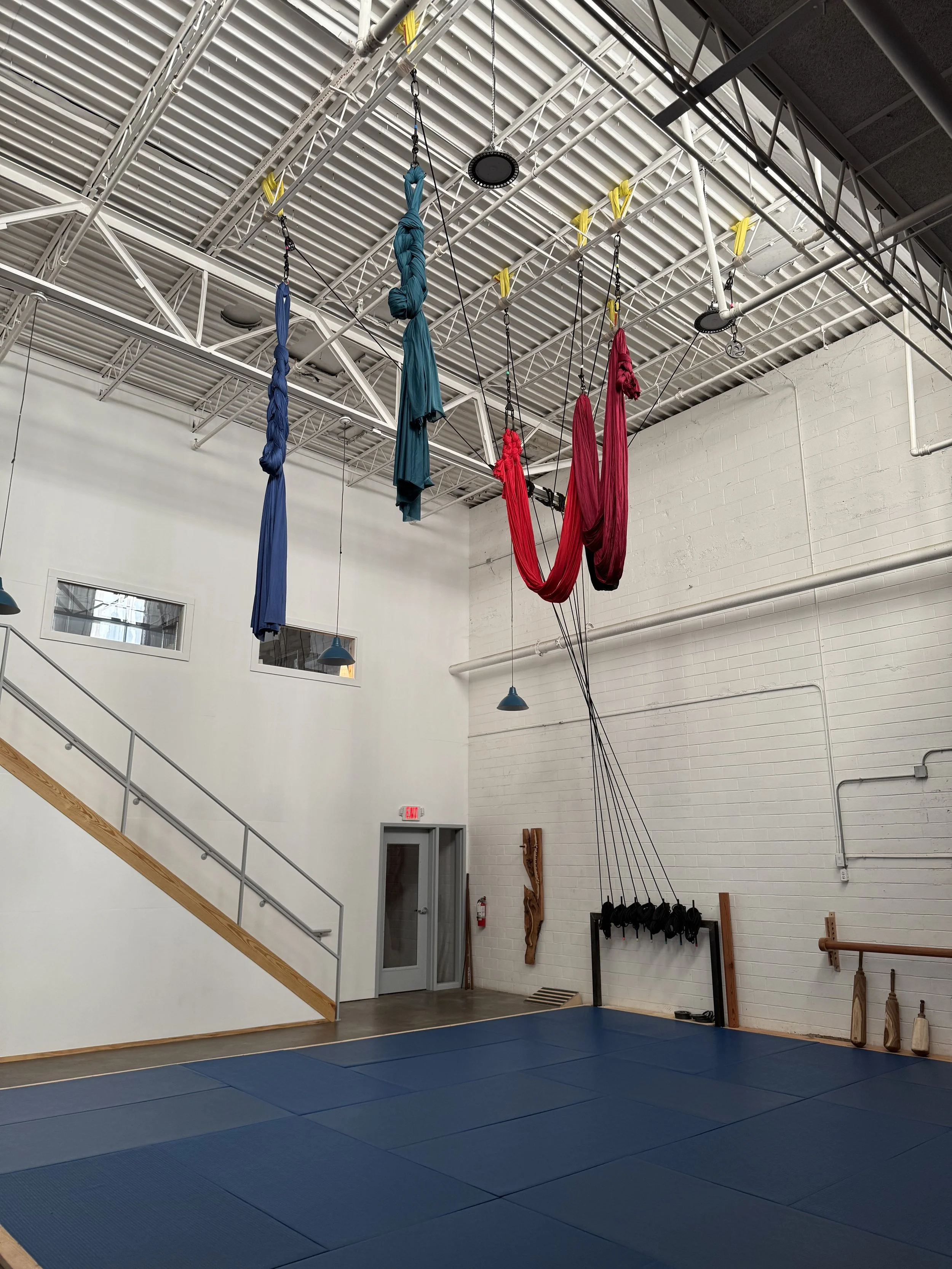 An indoor aerial fitness studio with colorful aerial silks hanging from the ceiling. The studio has a blue padded floor, white brick walls, and a staircase with a wooden handrail. There are overhead lights and a wall-mounted coat rack with hanging weights.