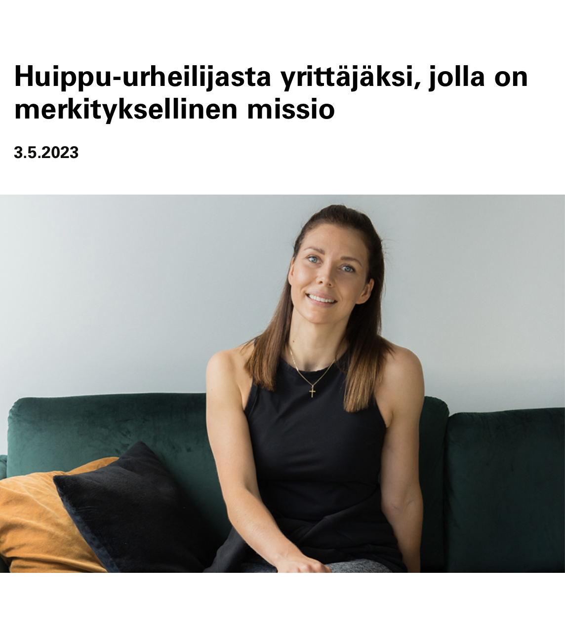 Nainen istuu vihreällä sohvalla, taustalla valkoinen seinä, päällään musta toppi ja kaulassa kultainen risti.