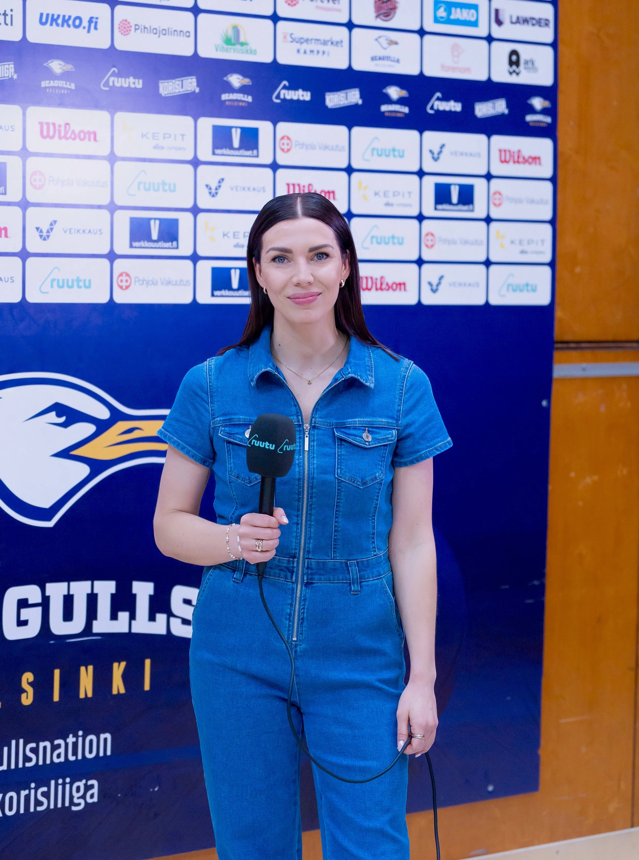 Nainen haastattelussa sisätiloissa, taustalla sponsorilappuja ja merkkejä, lukee mm. 'Gulls', 'Wilson', 'Kepit', 'verkkouutiset', 'ruutu', 'liiga'. Naisella tummat hiukset ja sininen farkkuasu, hän pitää mikrofonia ja puhelinta.