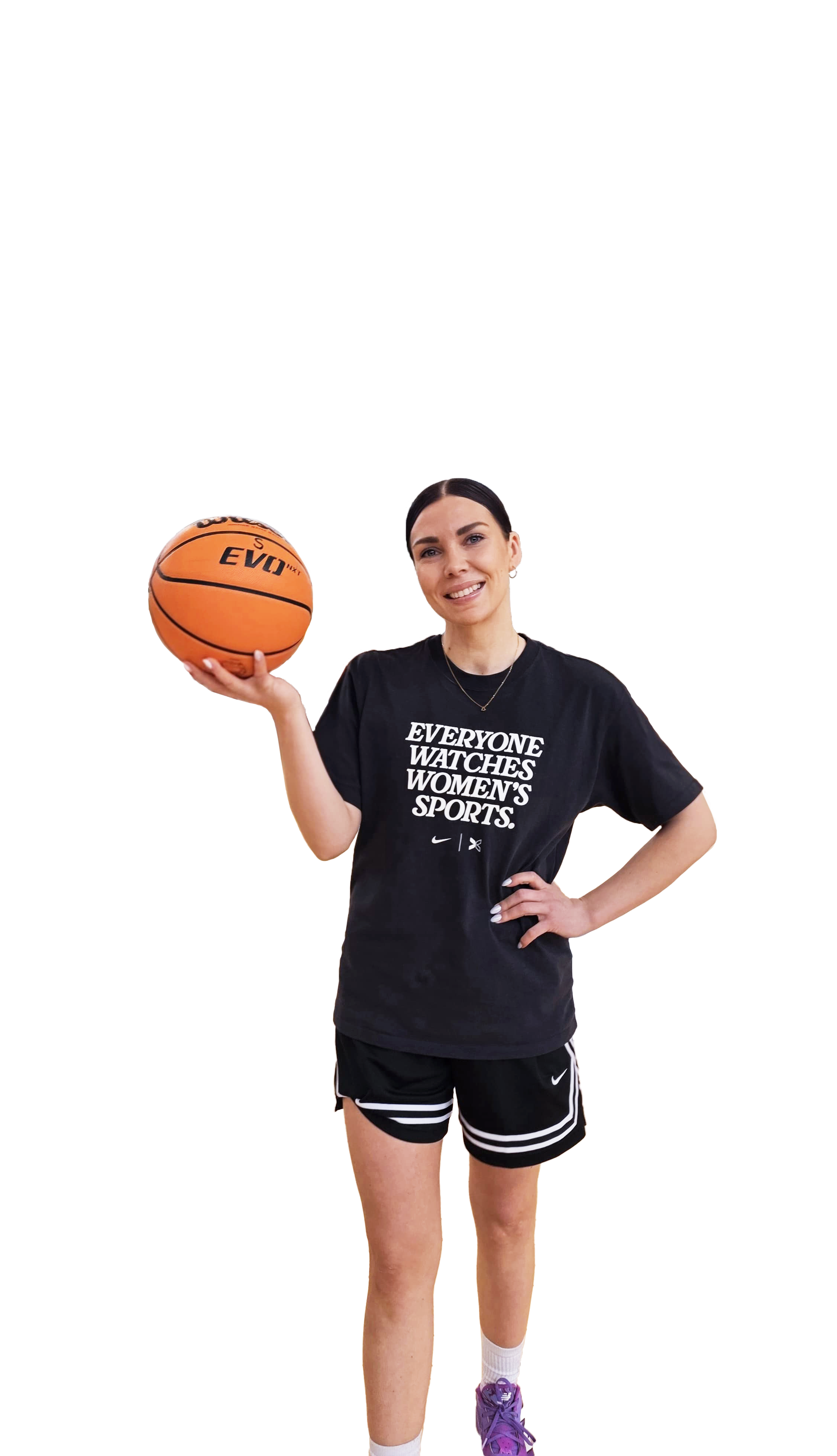 Nuori nainen pitelee koripalloa oikeassa kädessään ja hymyilee. Hänellä on musta t-paita, jossa lukee 'Everyone watches women's sports', sekä mustat shortsit ja purple-violetit urheilukengät.