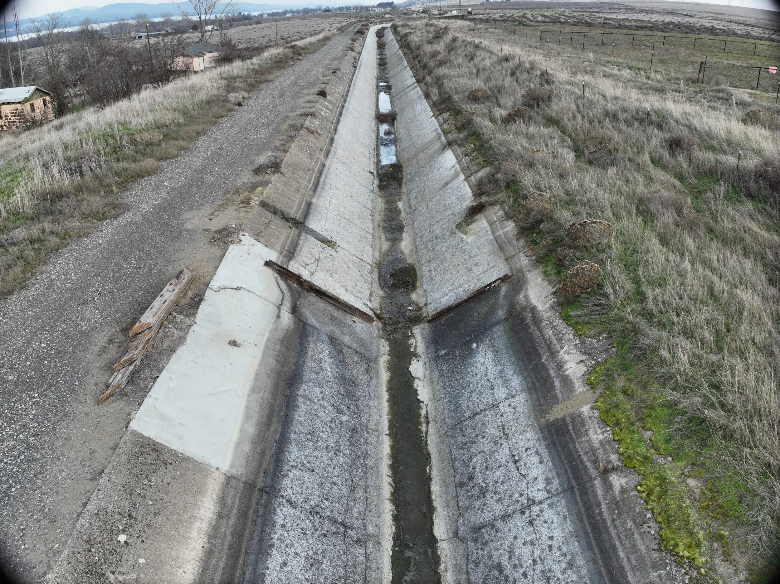 A dry, cracked, concrete drainage ditch running through a rural area with a dirt road on the left, some small structures on the left, and grassy fields on the right, with some mountains in the background.