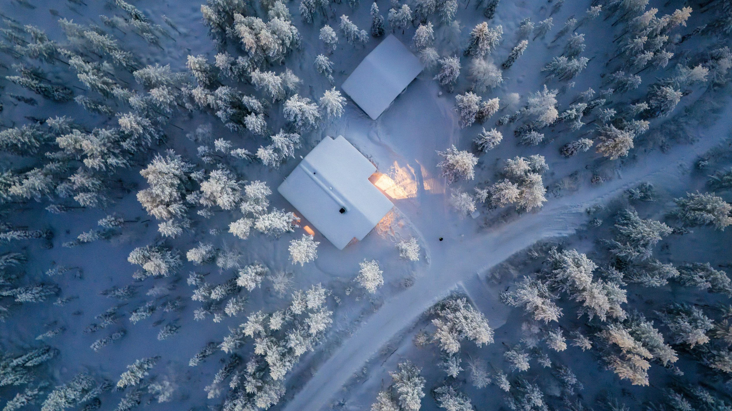 Aerial view of a snow-covered house and a smaller building among snow-covered trees on a winter day, with a lit fire inside.