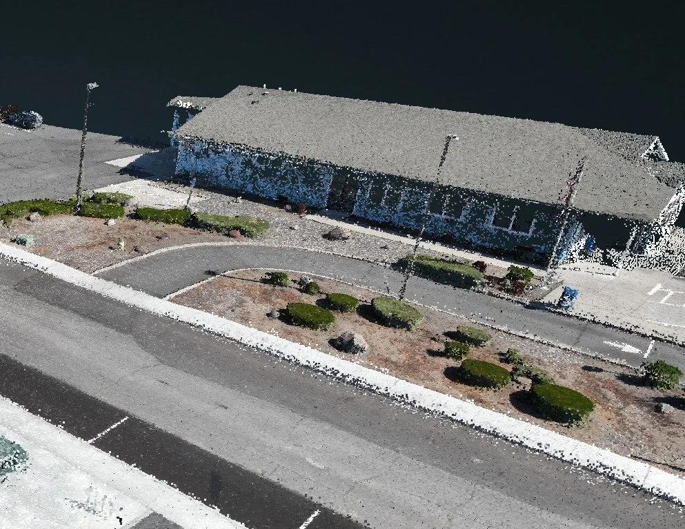An aerial view of a parking lot and a waterfront building with a sloped roof, surrounded by a sidewalk, street, and landscaped area with bushes.