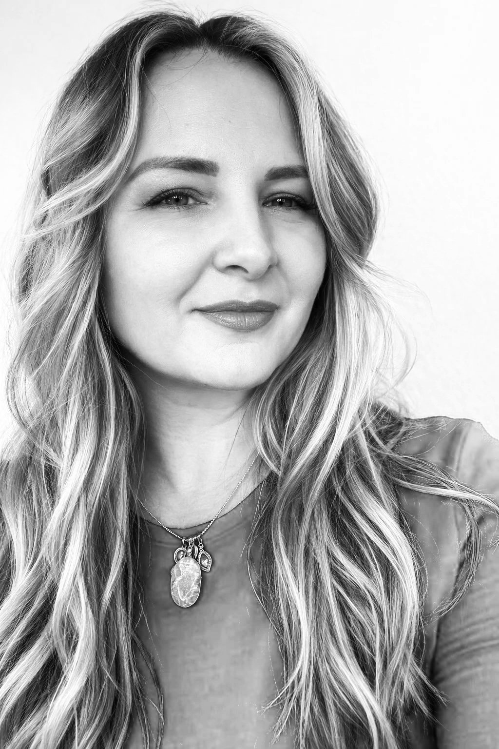 Black and white portrait of a woman with long wavy hair, wearing a necklace with a large pendant, smiling softly at the camera.