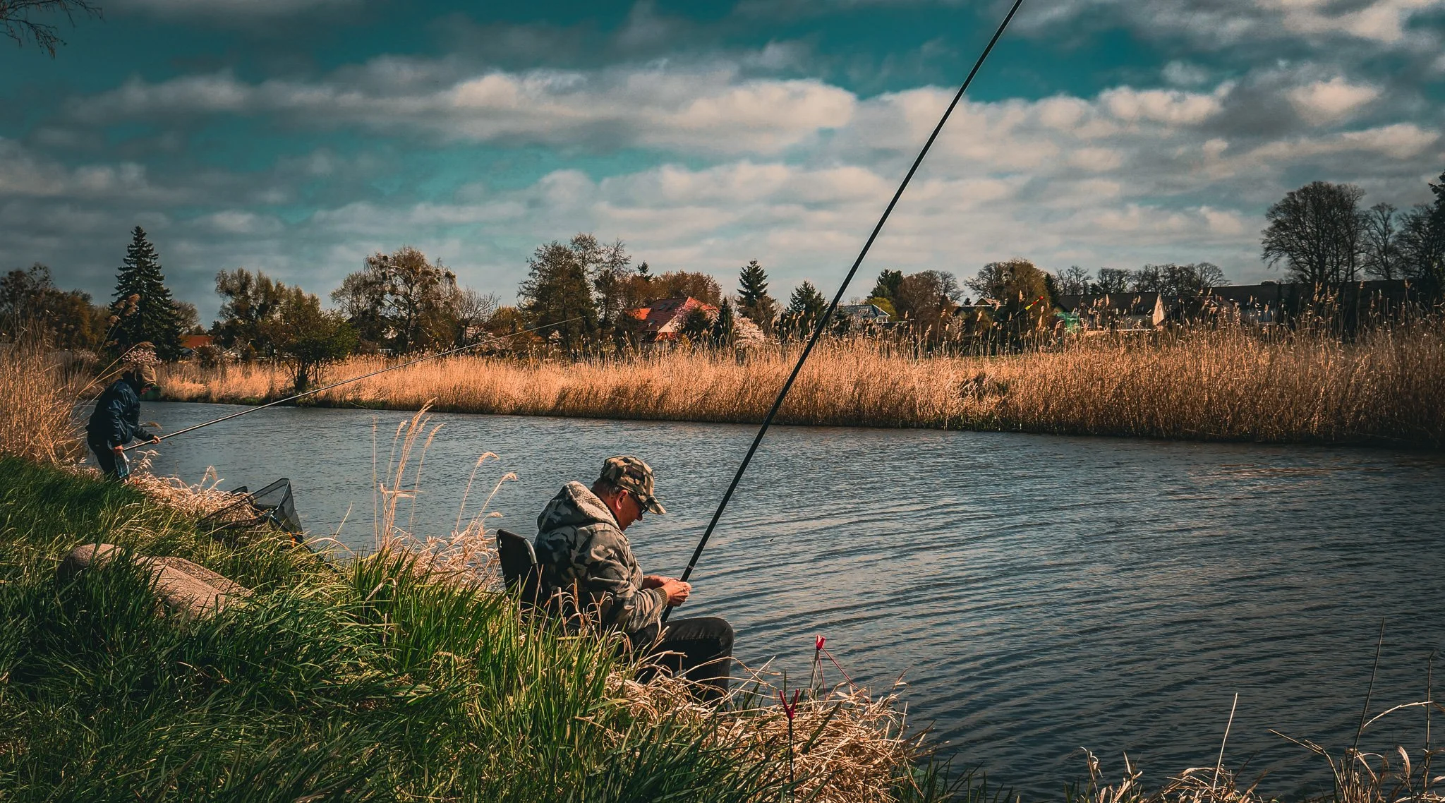 Zwei Angler sitzen an einem Flussufer und fischen, im Hintergrund sind Bäume, Häuser und ein bewölkter Himmel zu sehen.