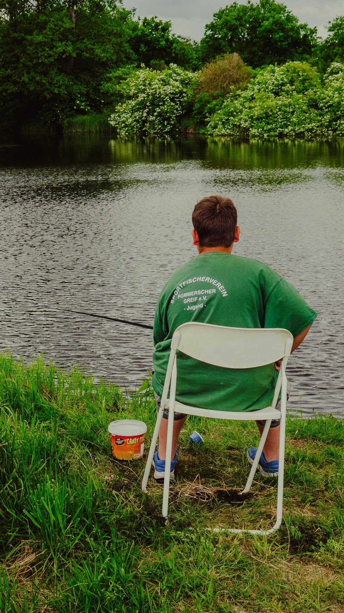 Mann sitzt auf einem weißen Klappstuhl am Ufer eines Gewässers und fischt. Er trägt ein grünes T-Shirt mit Aufdruck auf dem Rücken, und es gibt einen Eimer mit Candy Floss und eine Flasche Wasser auf dem Boden neben ihm, im Vordergrund grüne Gras.