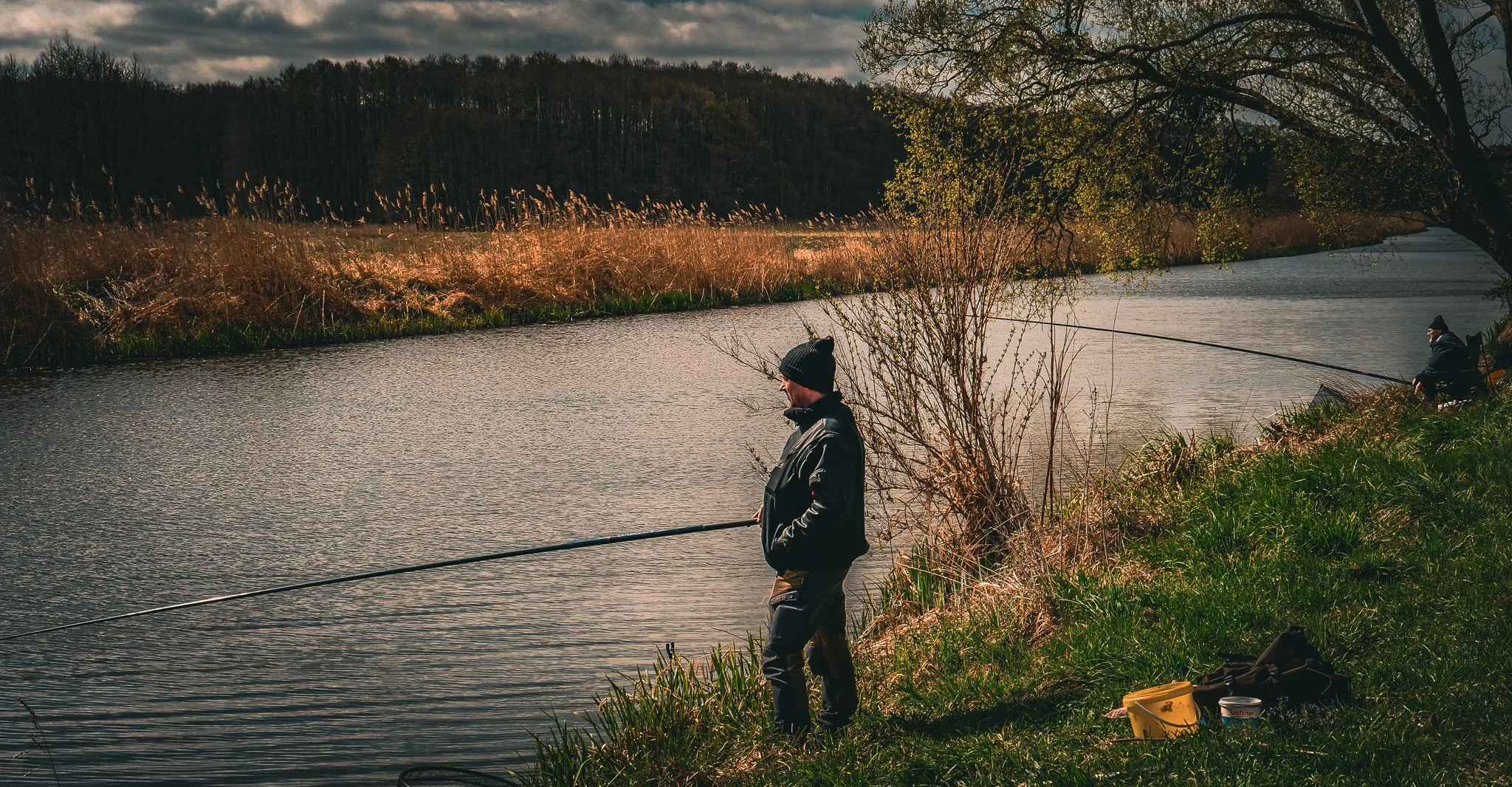 Zwei Männer beim Angeln an einem Fluss, umgeben von Gras und Bäumen, bei bewölktem Himmel.