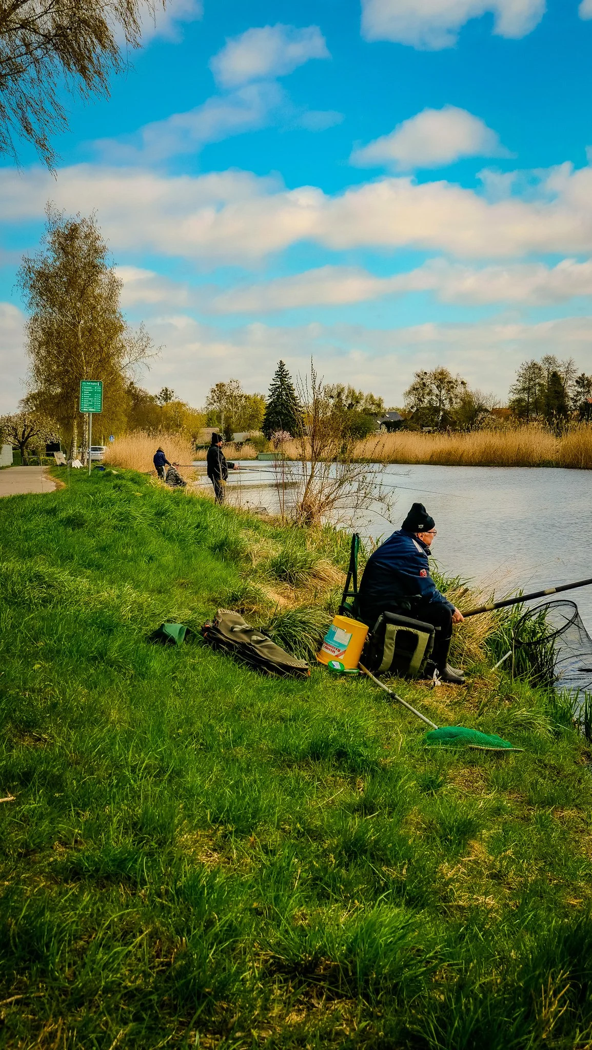 Menschen beim Angeln an einem Fluss unter einem blauen Himmel mit Wolken, umgeben von Bäumen und Gräsern
