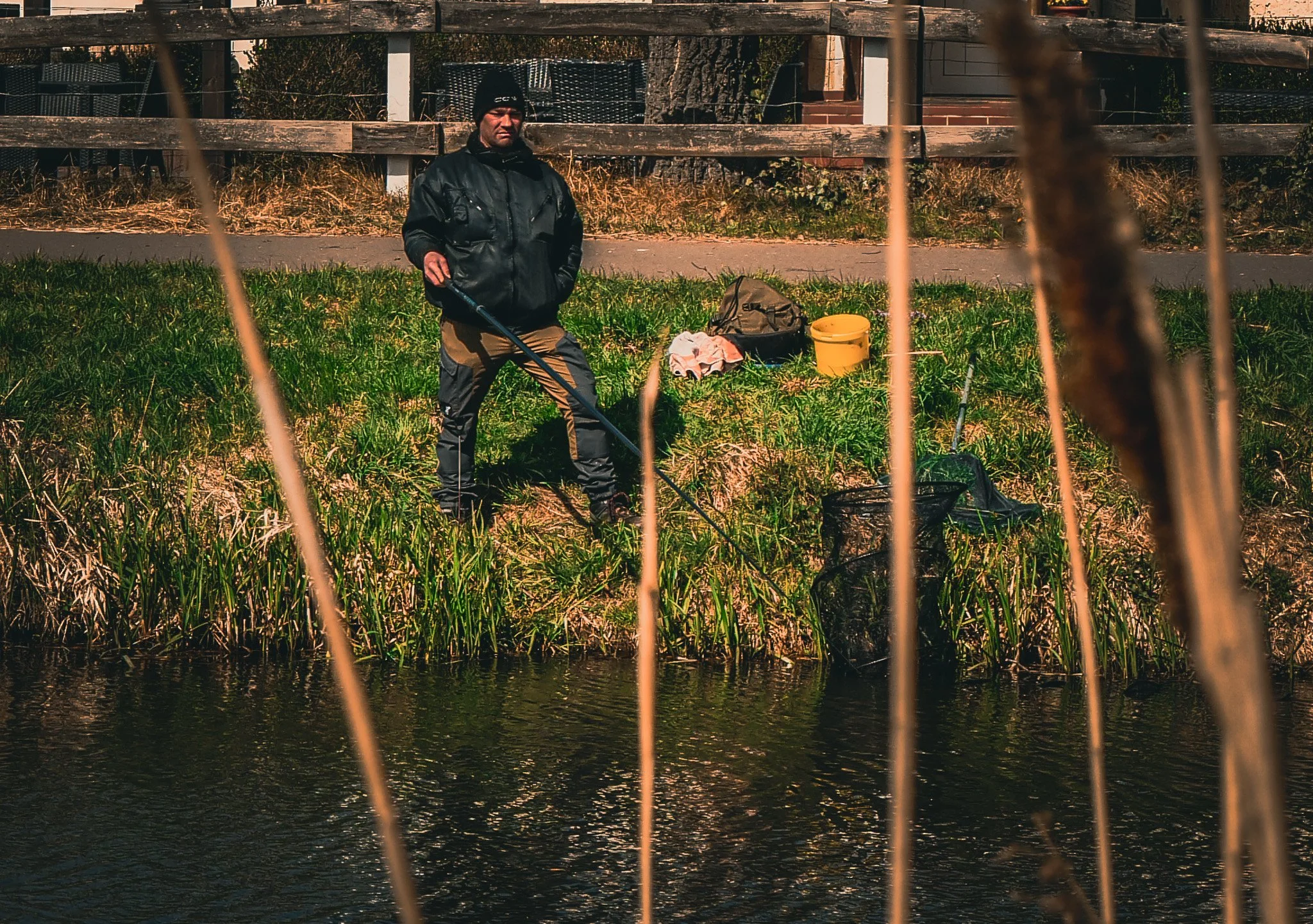 Ein Mann steht an einem Flussufer, blickt nach vorne, trägt eine schwarze Jacke und eine schwarze Mütze. Vor ihm sind Gräser, auf dem Boden liegen eine Tasche, eine Schirmmütze, ein gelber Eimer und andere Gegenstände. Im Hintergrund befinden sich ei