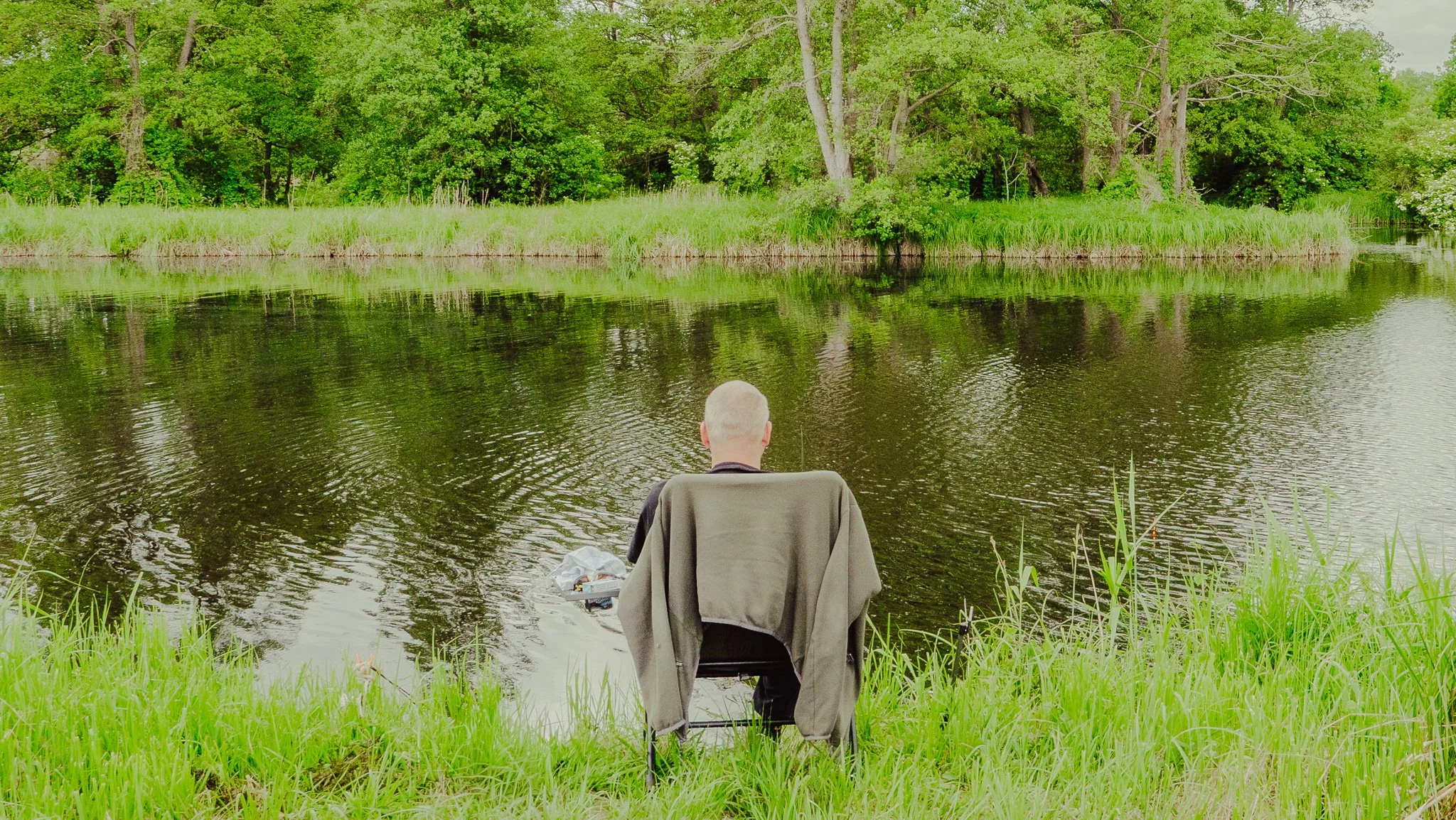 Ein Mann sitzt an einem Ufer eines Flusses, umgeben von grünen Bäumen und Gras, beim Angeln.