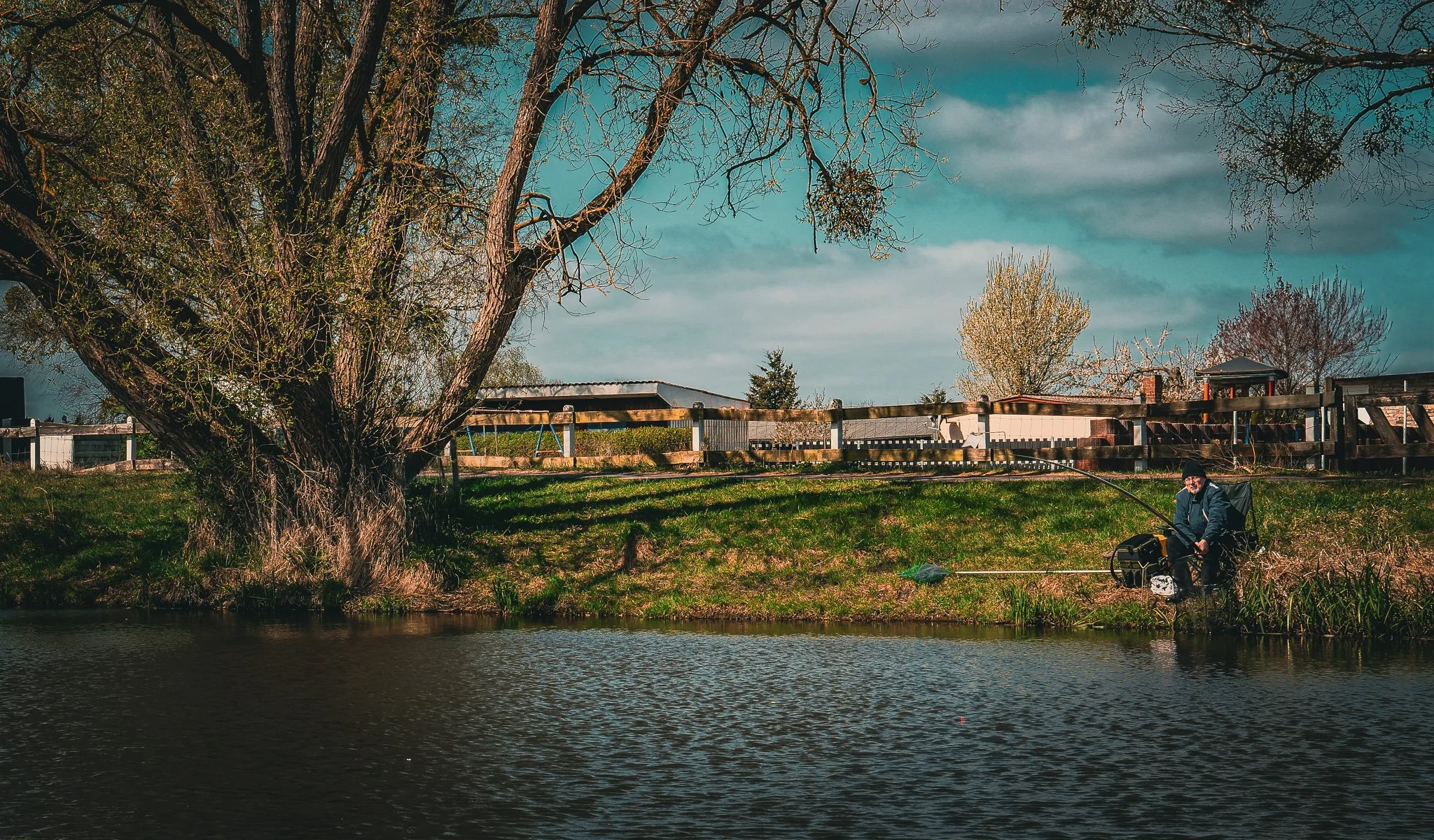 Ein Mann sitzt am Ufer eines Flusses, im Hintergrund Bäume, eine Holzzäune, und ein Haus. Er ist beim Angeln mit einer Angelschnur und einem Angelhaken. Es ist Frühlings- oder Herbstdämmerung mit bewölktem Himmel.