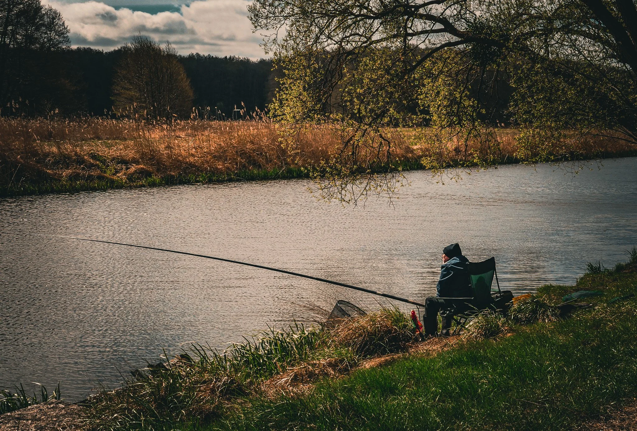 Junge Person angelt an einem Fluss, umgeben von Büschen und Bäumen, bei bewölktem Himmel.