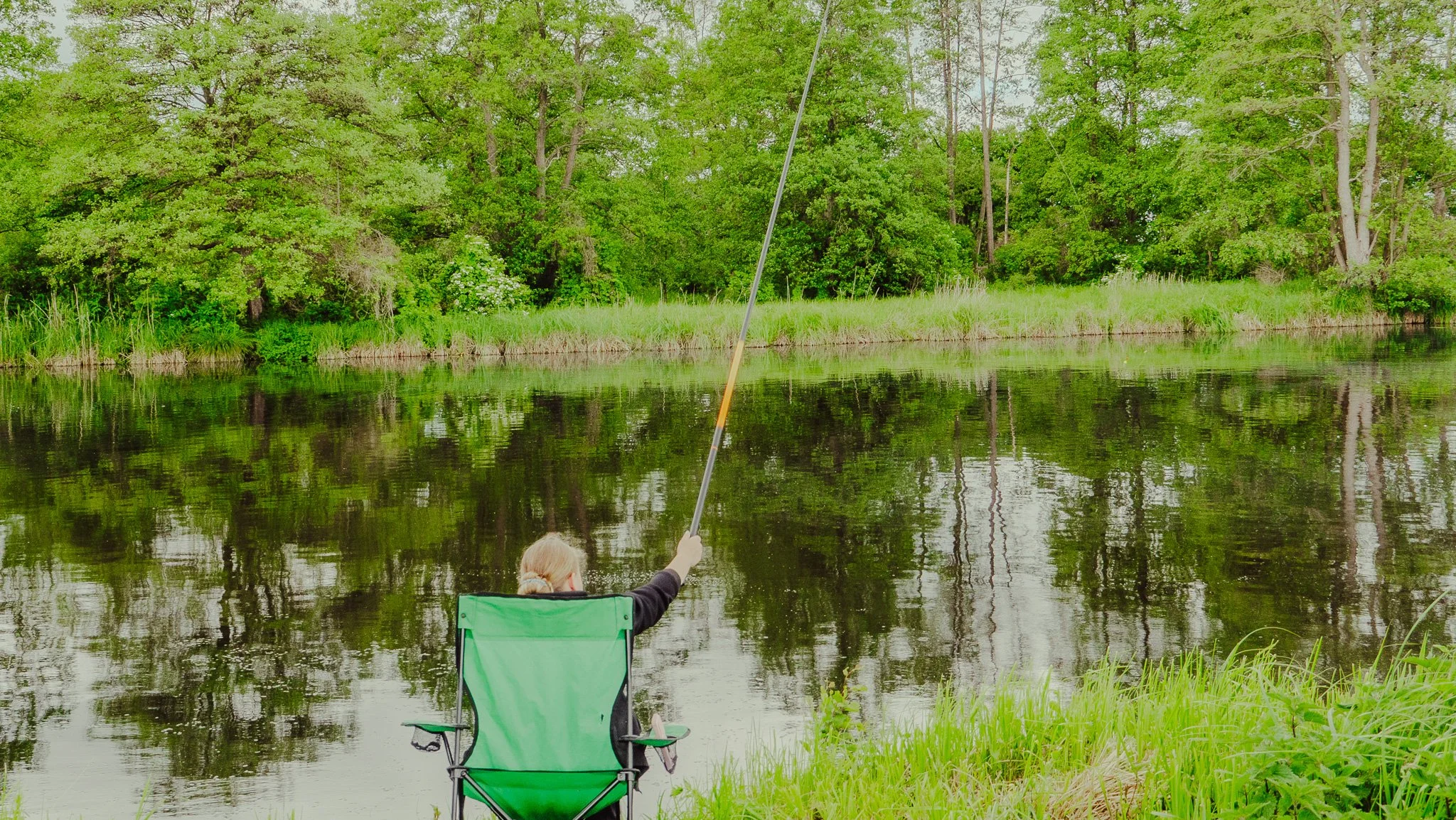 Ein Kind sitzt in einem grünen Campingstuhl am Ufer eines Sees und fischt mit einer Angel. Der Hintergrund zeigt grüne Bäume und laubgesäumte Wasserflächen.