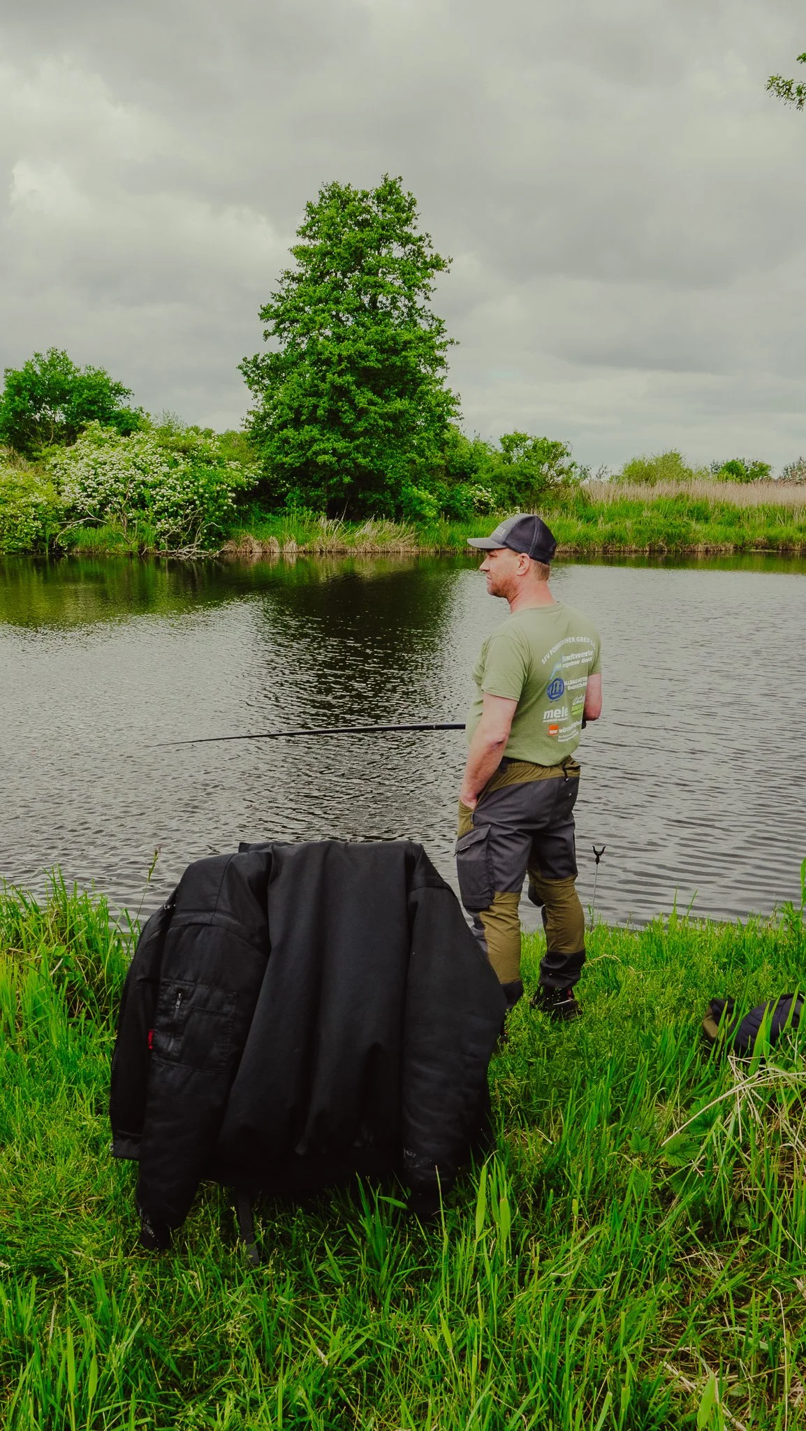 Mann beim Angeln am Fluss unter einem bewölkten Himmel, mit Rucksack und Angelzubehör im Gras.