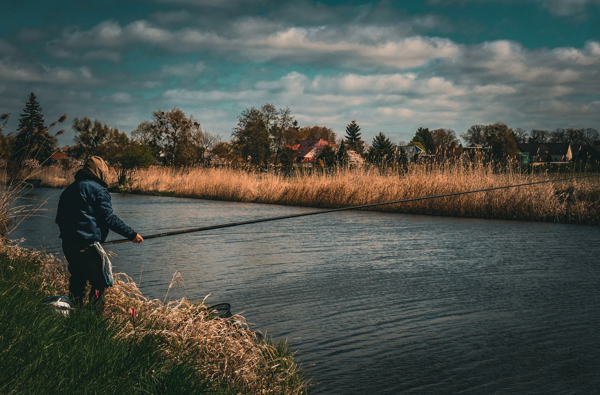 Eine Person angelt an einem Fluss, umgeben von hohen Gräsern und Bäumen, mit Häusern im Hintergrund bei bewölktem Himmel.