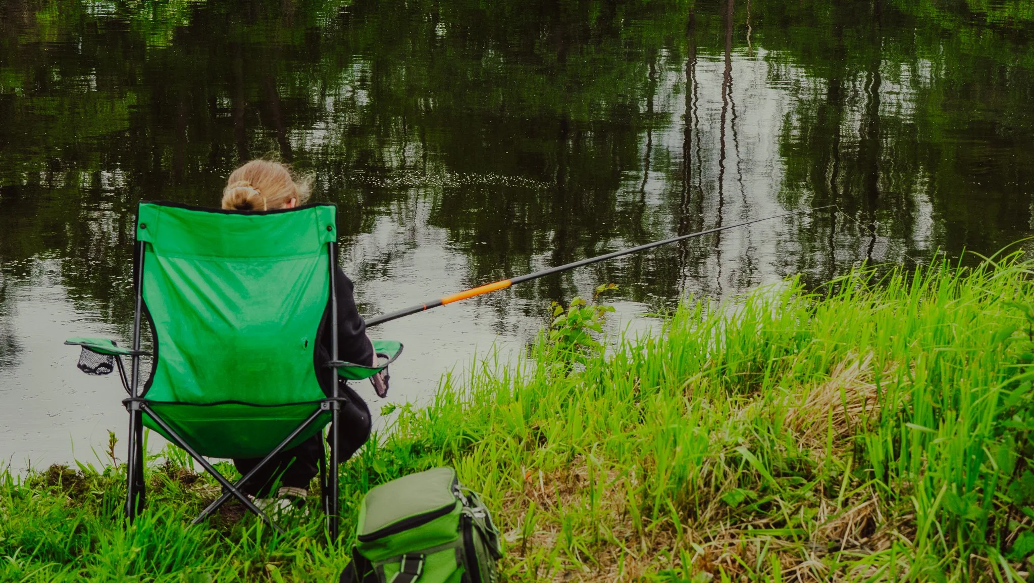 Eine Person sitzt in einem grünen Klappstuhl am Ufer eines Flusses und fischt mit einer Angel.