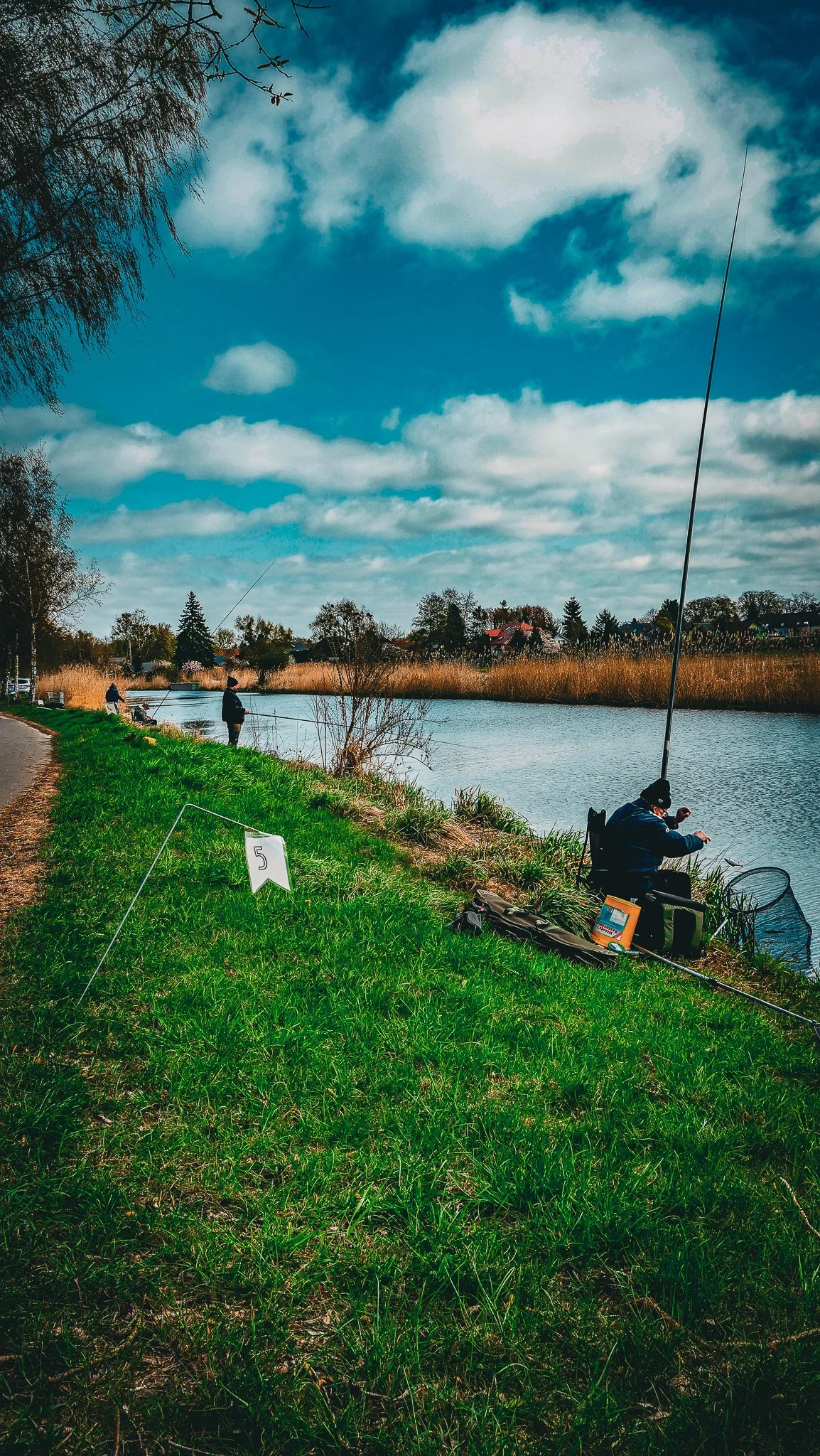 Menschen beim Angeln an einem Fluss, mit einem grünen Ufer, eine Straße und Häuser im Hintergrund, bewölkter Himmel.