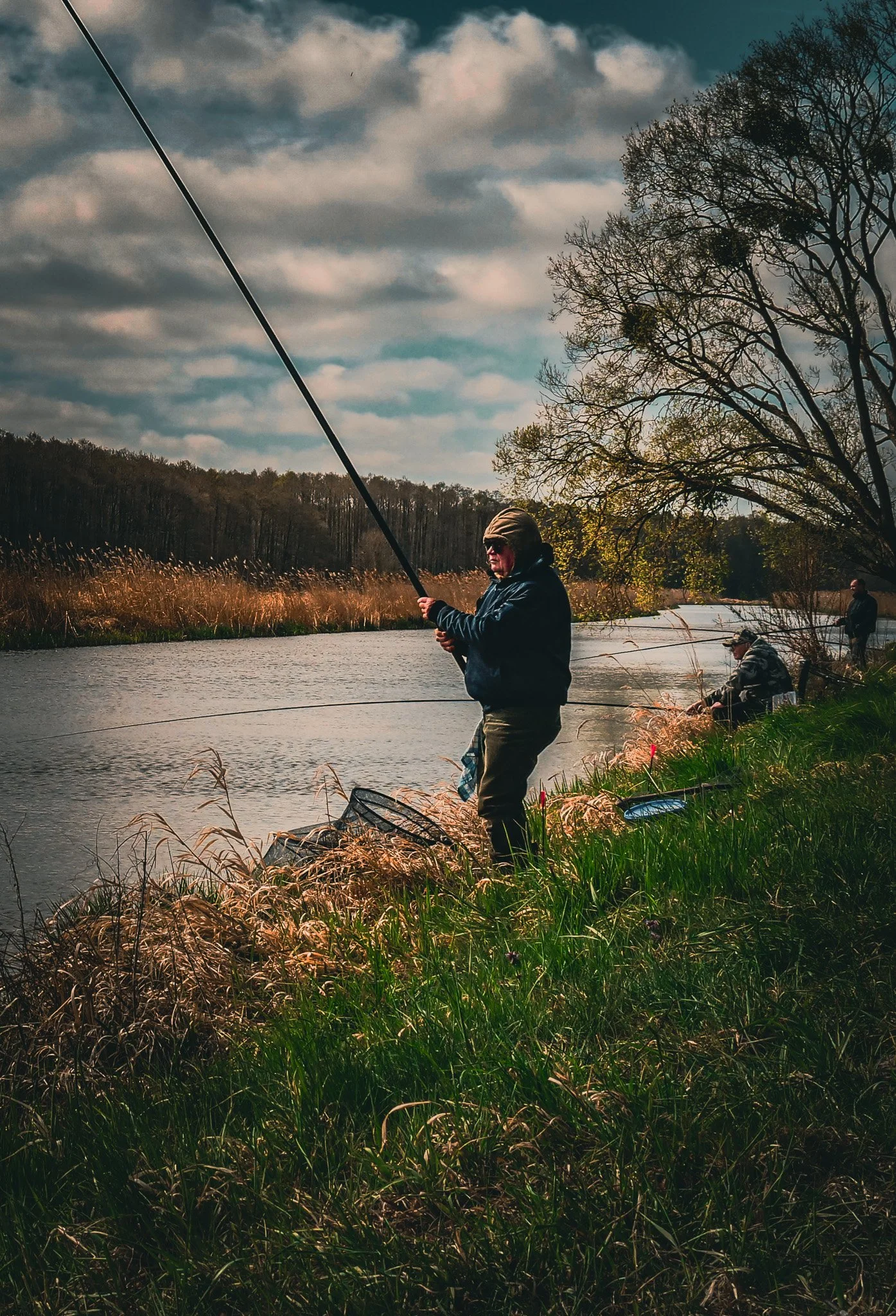 Fischer steht am Ufer eines Flusses, hält eine Angelrute und versucht, einen Fisch zu fangen, während andere am Flussufer sitzen und angeln. Die Umgebung ist von Bäumen umgeben, der Himmel ist bewölkt.