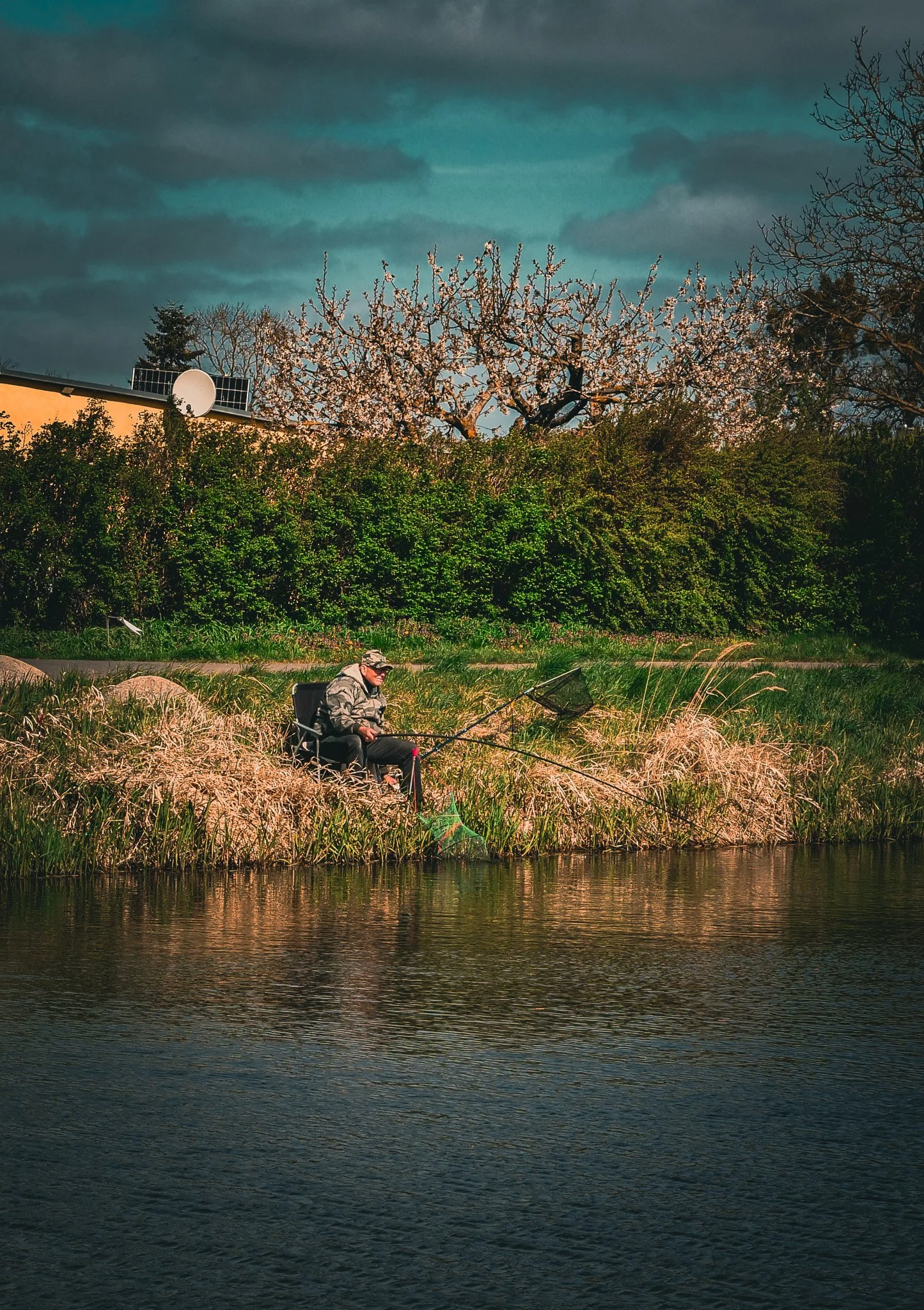 Ein Mann sitzt am Ufer eines Flusses und fischt mit einer Angel. Im Hintergrund sind Bäume, einige blühen, und das Wetter ist bewölkt.
