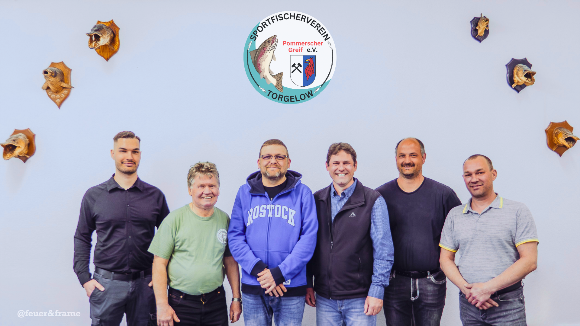 Gruppenfoto von sechs Männern, die vor einer weißen Wand stehen, mit einem Vereinswappen des Sportfischervereins Pommerscher Greif e.V. Torgelow und gehängten Wildkatzen- und Fischschnitzereien