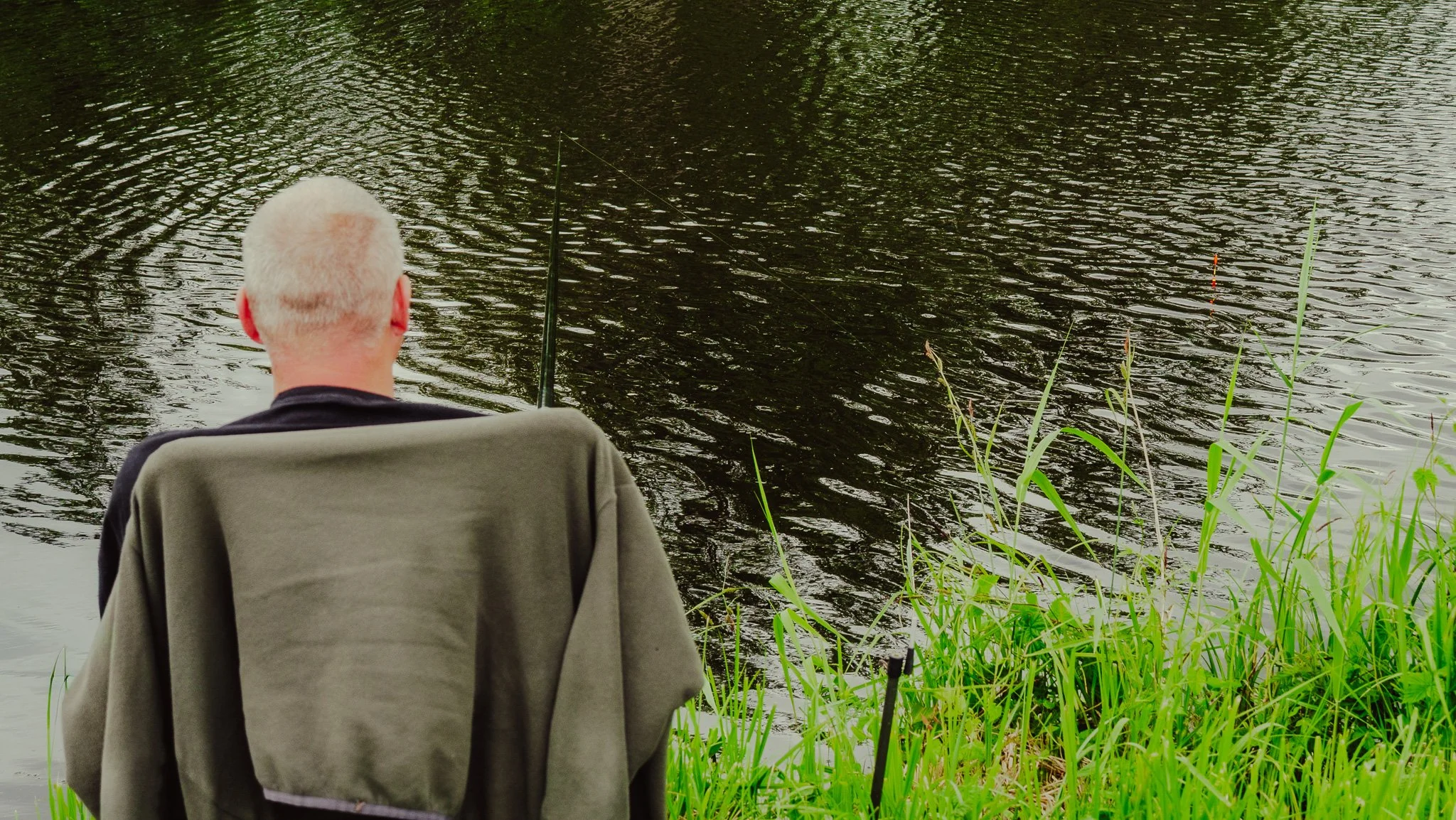 Ein älterer Mann sitzt im Freien am Wasser, umgeben von Gräsern, beim Angeln.