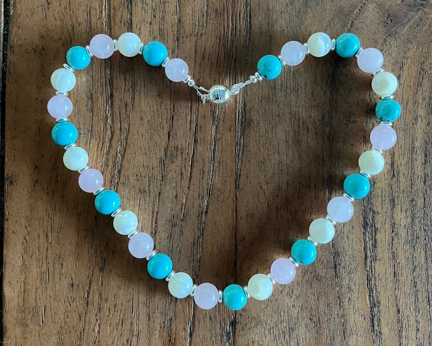 A beaded necklace made with alternating turquoise, white, and light purple beads, laid out on a wooden surface.