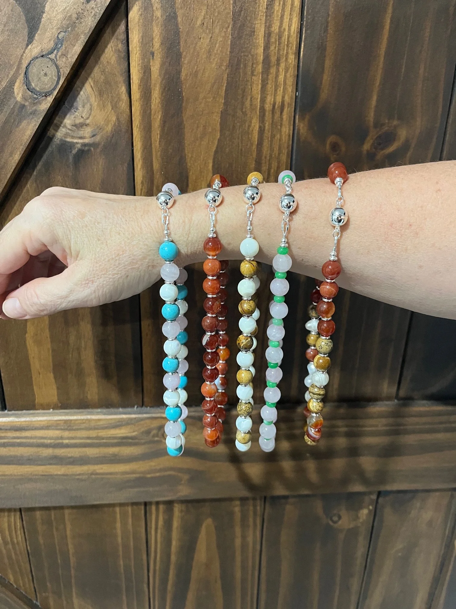 Several beaded necklaces hanging from a person's wrist, with each necklace made of multicolored gemstone beads with magnetic metal clasps, against a wooden background.