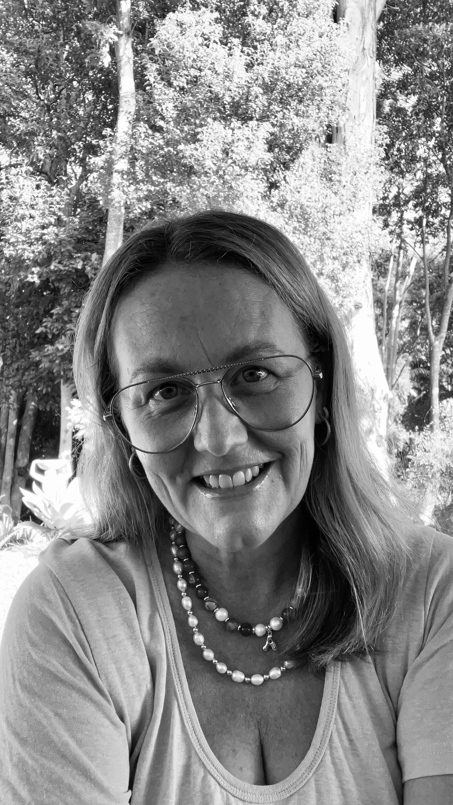 A woman with glasses smiling outdoors with trees and sunlight in the background.