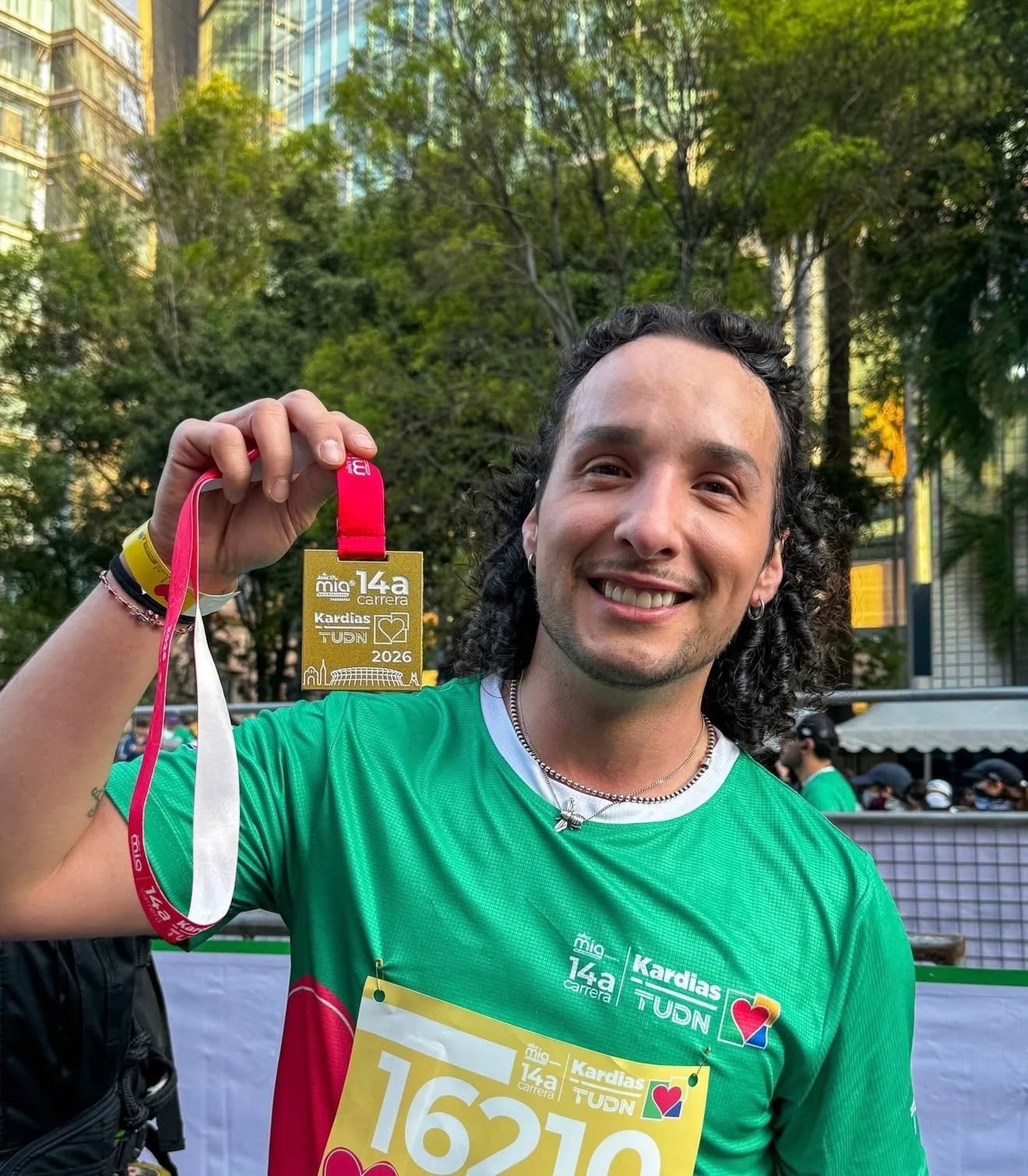 Hombre con cabello rizado y camiseta verde, sonriendo, sosteniendo una medalla dorada de la carrera 14a. presente en el evento de la carrera de Kardias TUDN en 2026, en un entorno urbano con árboles y edificios de fondo.
