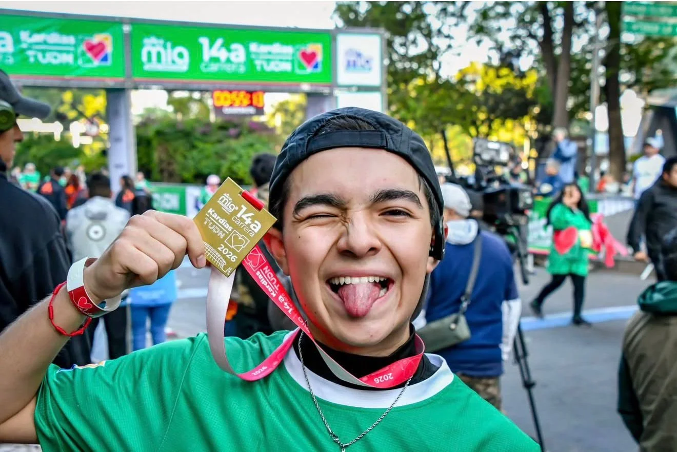 Persona joven con camiseta verde, gorra negra, mostrando un certificado y una medalla de la carrera 'Mío 14a' en un evento al aire libre con muchas personas y cámaras en el fondo.