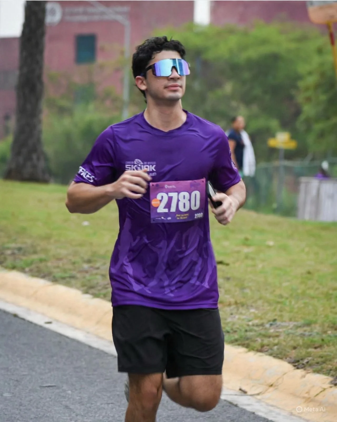 Joven corriendo en una carrera, lleva una camiseta púrpura, número 2780, gafas reflectantes azules, y sostiene un teléfono móvil en la mano derecha. Está en un entorno al aire libre con césped y árboles.