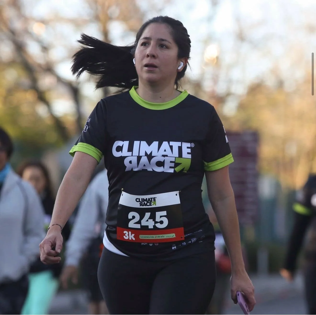 Una mujer corriendo en una carrera, lleva una camiseta negra con detalles verdes y el texto 'CLIMATE RACE'. Tiene el número 245 y una etiqueta que indica la distancia '3k'.
