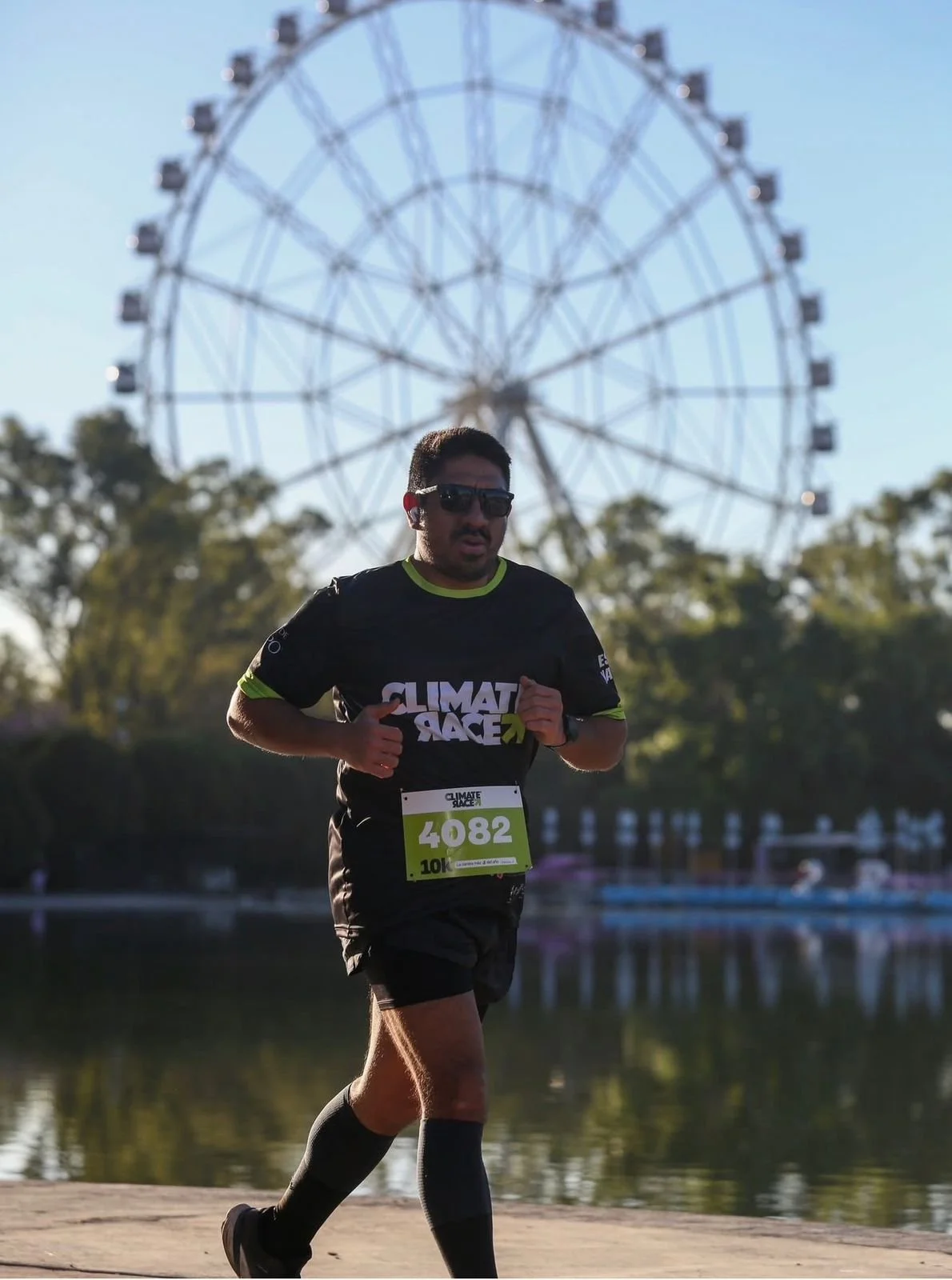Hombre corriendo en una maratón en un parque con una rueda de feria grande al fondo. Lleva gafas de sol, camiseta negra con logo de 'CLIMATE RACE', número de participante 4082, y ropa deportiva.