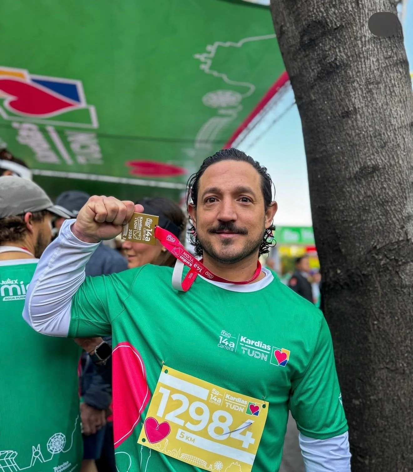 Hombre con barba y bigote sosteniendo una medalla en una carrera. Lleva una camiseta verde con el número 12984 y un pase de corredor. Está en un evento de maratón con otras personas y un árbol cerca.